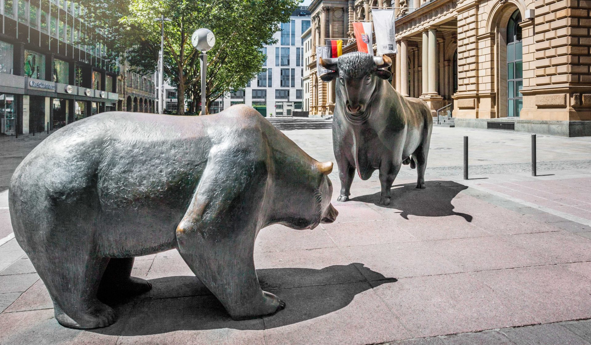 Das Bild zeigt die berühmten Skulpturen von einem Bär und einem Stier vor dem Gebäude der Frankfurter Börse in Frankfurt am Main, Deutschland. Diese Skulpturen repräsentieren den dynamischen Charakter der Finanzmärkte. 

**Symbolik der Darstellungen:**

1. **Bär:**
   - **Bedeutung:** Der Bär steht für einen "Bärenmarkt," d.h., eine Phase rückläufiger oder stagnierender Aktienkurse.
   - **Merkmal:** Ein Bärenmarkt wird durch Pessimismus, sinkende Preise und negative Erwartungen charakterisiert.

2. **Stier:**
   - **Bedeutung:** Der Stier symbolisiert einen "Bullenmarkt," d.h., eine Phase steigender Aktienkurse.
   - **Merkmal:** Er ist durch Optimismus, steigende Preise und positive Zukunftserwartungen gekennzeichnet.

**Standort:** Diese Skulpturen sind ikonische Wahrzeichen, die auf dem Börsenplatz vor dem historischen Gebäude der Frankfurter Börse stehen. Besucher aus aller Welt kommen vorbei, um die Statuen zu fotografieren und die wirtschaftliche Atmosphäre zu erleben. 

Die Kombination dieser beiden Tiere steht für die ständige Wechselwirkung und Dynamik der globalen Finanzmärkte und spiegelt die vielfältigen Meinungen und Erwartungen der Marktteilnehmer wider.