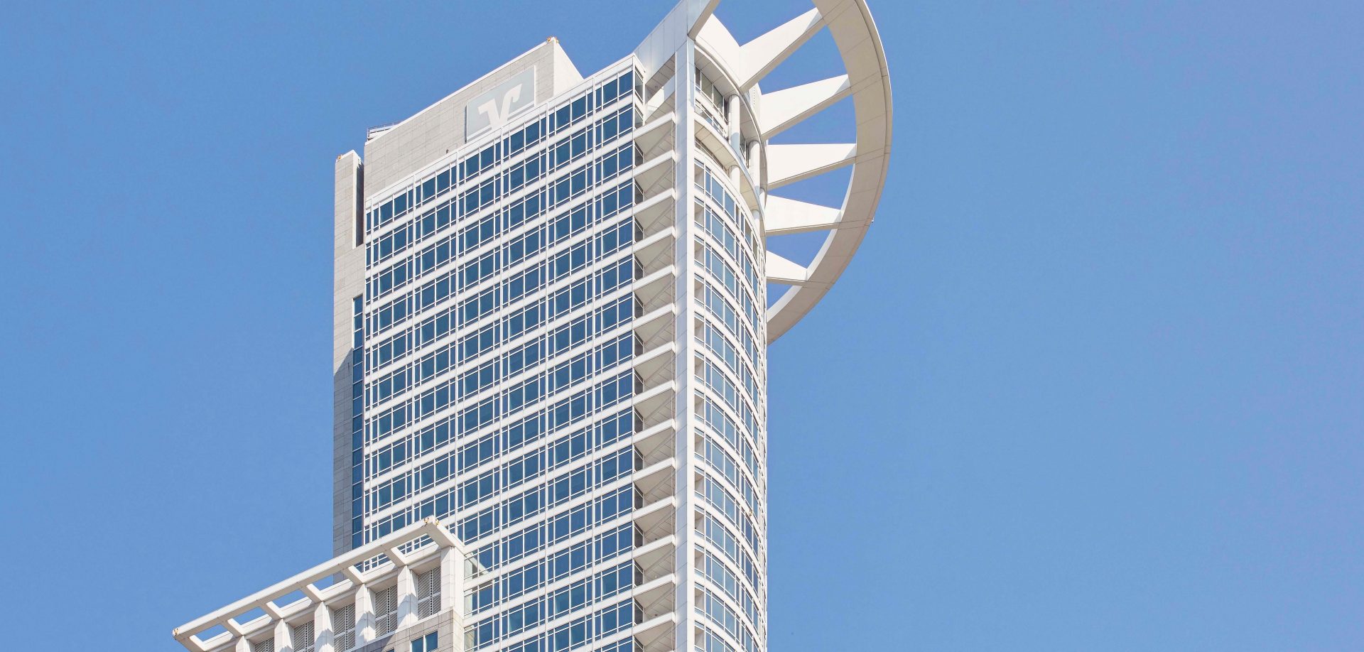 Das Bild zeigt ein modernes Hochhaus, das aufgrund seiner architektonischen Merkmale leicht zu erkennen ist. Die Struktur hebt sich durch das markante oberste Element ab – einen halbrunden Abschluss, der als Sonnenschutz oder rein dekoratives Element fungieren könnte.

**Architektonische Merkmale:**

1. **Glasfassade**: Die Gebäudehülle besteht überwiegend aus Glas, was die Verwendung von natürlichem Licht maximiert und eine hochmoderne Ästhetik verleiht.

2. **Kanten und Winkel**: Klar definierte Kanten und rechtwinklige Elemente geben dem Gebäude ein zeitgemäßes und geometrisches Aussehen.

3. **Oberes Element**: Der Halbkreis auf der Spitze des Gebäudes könnte entweder eine funktionale Rolle als strukturelles oder klimatisches Element haben oder einfach als Teil eines einzigartigen Designs.

**Möglicher Standort:**
- Solche architektonischen Stile sind häufig in Metropolen wie Singapur, Dubai oder Hong Kong zu finden, wo moderne Architektur floriert.

**Zusammenfassung:**
Das Bild zeigt ein Hochhaus, das ein Beispiel für zeitgenössische moderne Architektur ist, erkennbar durch seine Glasfassade und markante Spitzenstruktur. Es steht vermutlich in einer urbanen Umgebung und ist ein Ausdruck von Innovation und Modernität in der Bauweise.