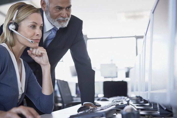 Das Bild zeigt eine Frau mit Headset und einen Mann im Anzug, die zusammen an einem Computerarbeitsplatz arbeiten. Die Szene wirkt wie ein typischer Büro- oder Callcenter-Arbeitsplatz, und beide Personen schauen konzentriert auf den Bildschirm.