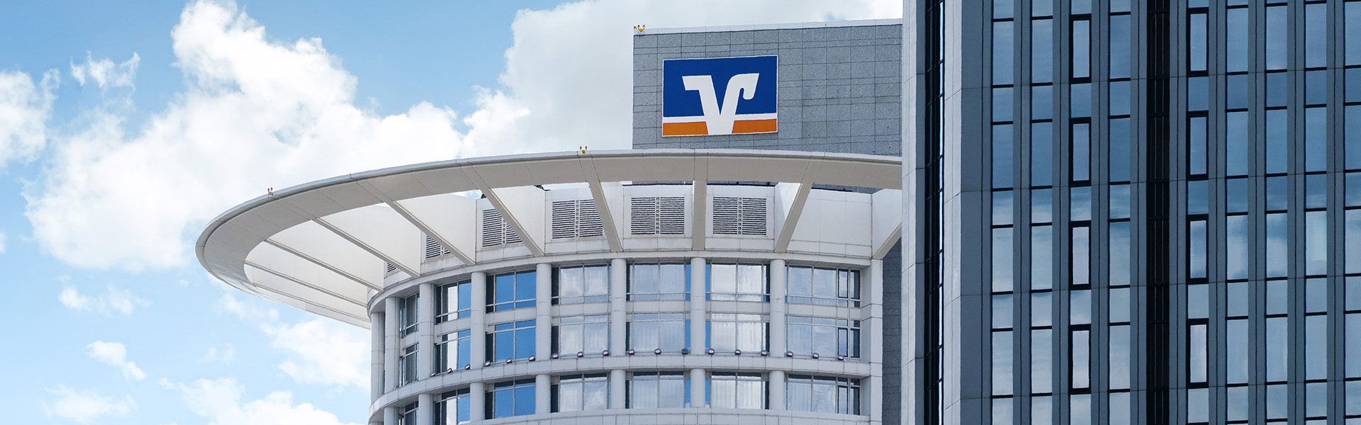 Das Bild zeigt ein Gebäude mit dem Logo der Volksbank. Es ist ein modernes Bauwerk mit einer runden Struktur und mehreren Fenstern.