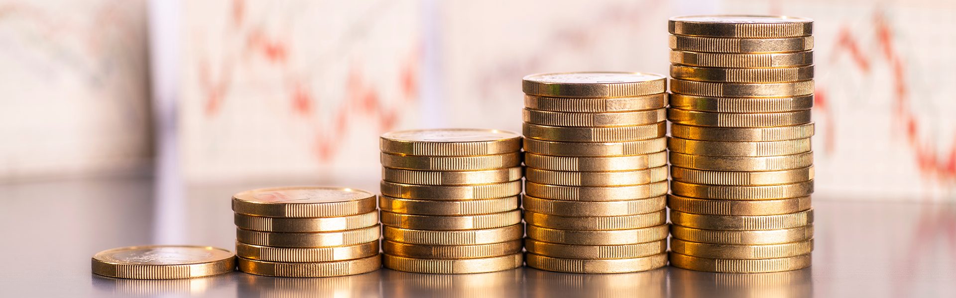 Das Bild zeigt gestapelte Münzen in steigender Höhe, die auf einem Tisch liegen. Im Hintergrund sind unscharfe Diagramme oder Charts zu erkennen, was auf finanzielle Themen wie Wachstum oder Investitionen hinweisen könnte.