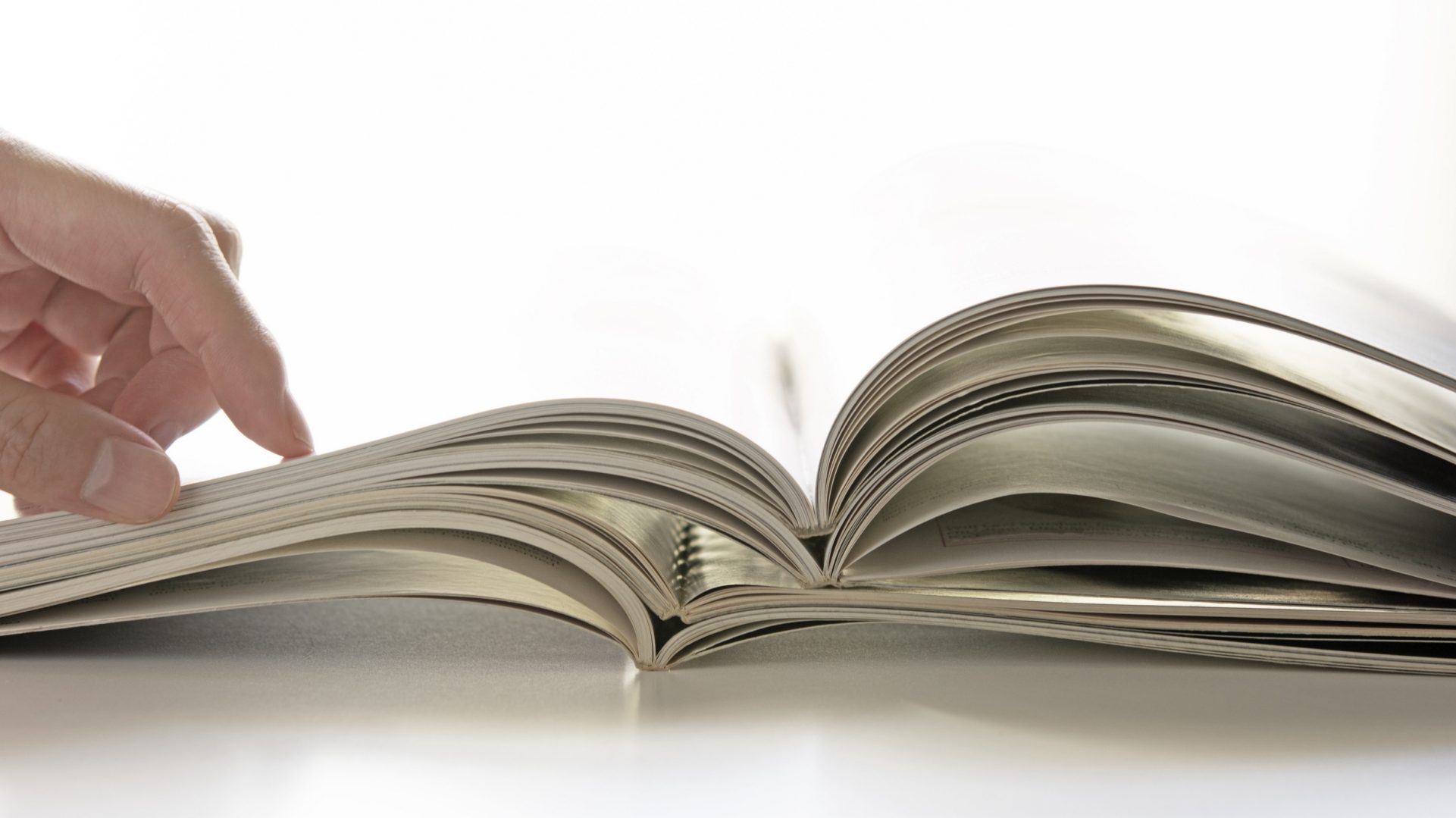 Das Bild zeigt ein geöffnetes Buch, wobei eine Hand dabei ist, durch die Seiten zu blättern. Die Perspektive vermittelt das Gefühl, durch das Buch zu schauen, möglicherweise in einem Moment des Lesens oder Durchstöberns. Das Buch liegt auf einer weißen Oberfläche, die das Licht reflektiert und eine helle Umgebung erzeugt.
