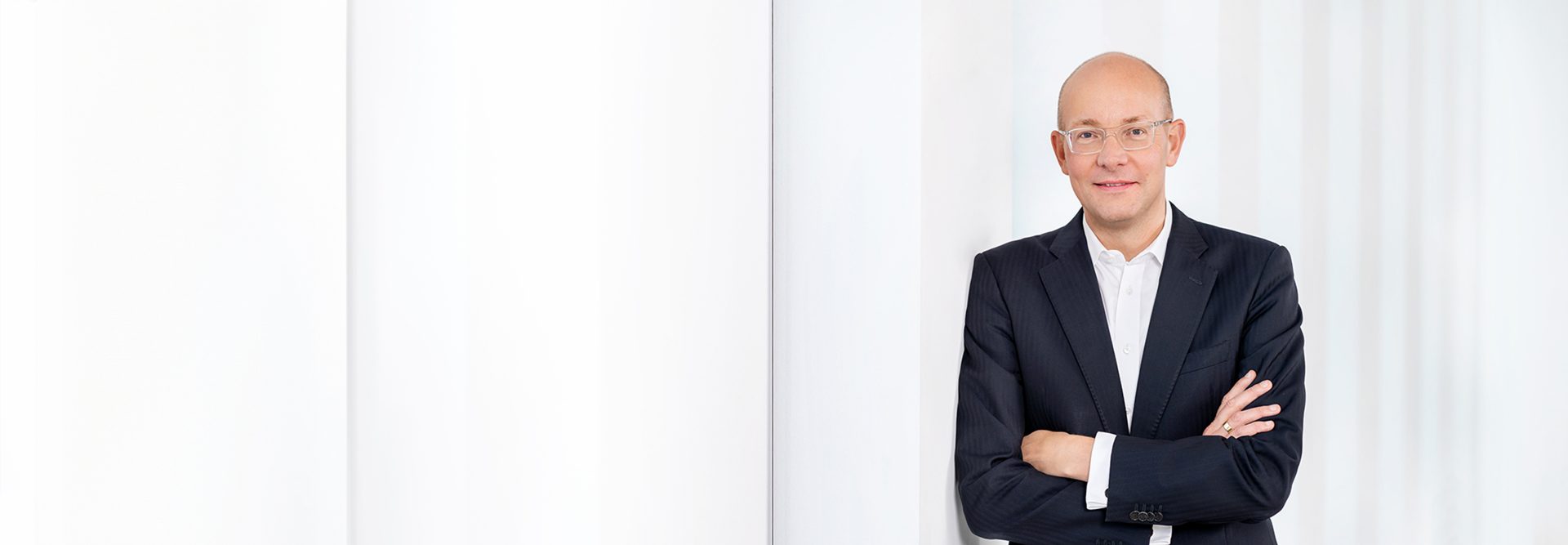 Das Bild zeigt einen Mann im Anzug, der mit verschränkten Armen vor einem hellen, modernen Hintergrund steht. Der Mann ist glatzköpfig und trägt eine Brille. Es handelt sich um ein professionell aufgenommenes Porträtfoto.