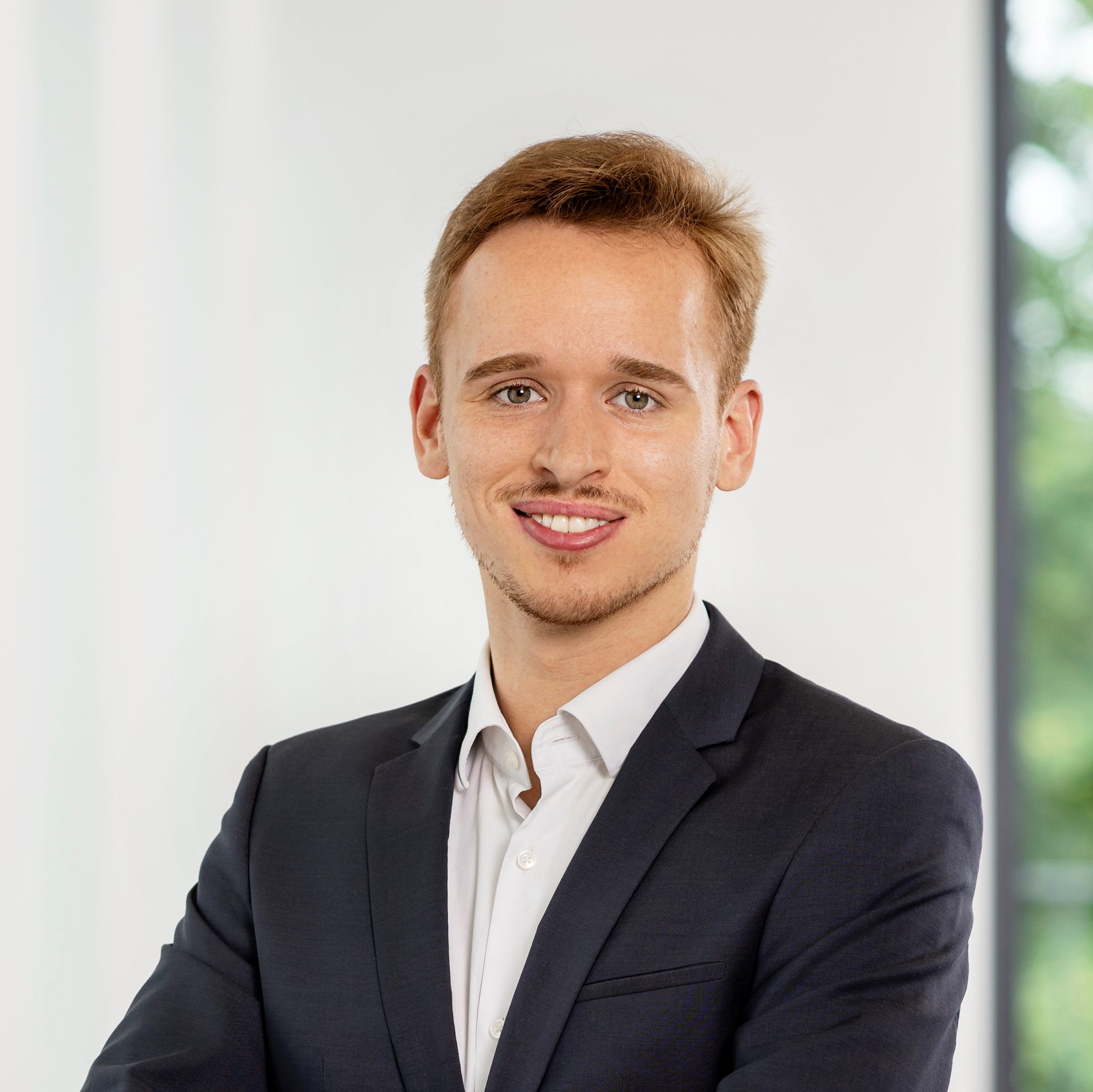 Die abgebildete Person trägt einen hellen Business-Anzug mit einem weißem Hemd. Der Hintergrund scheint ein helles Interieur mit einem großen Fenster zu sein, durch das etwas Grün von draußen sichtbar ist.