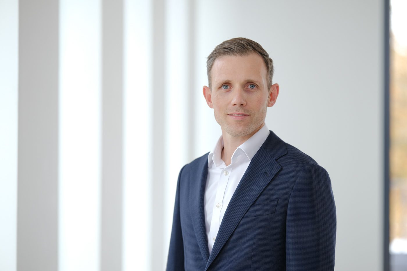 Eine Person in einem dunklen Anzug und weißem Hemd steht in einem hellen Raum mit einer Reihe von Fenstern.