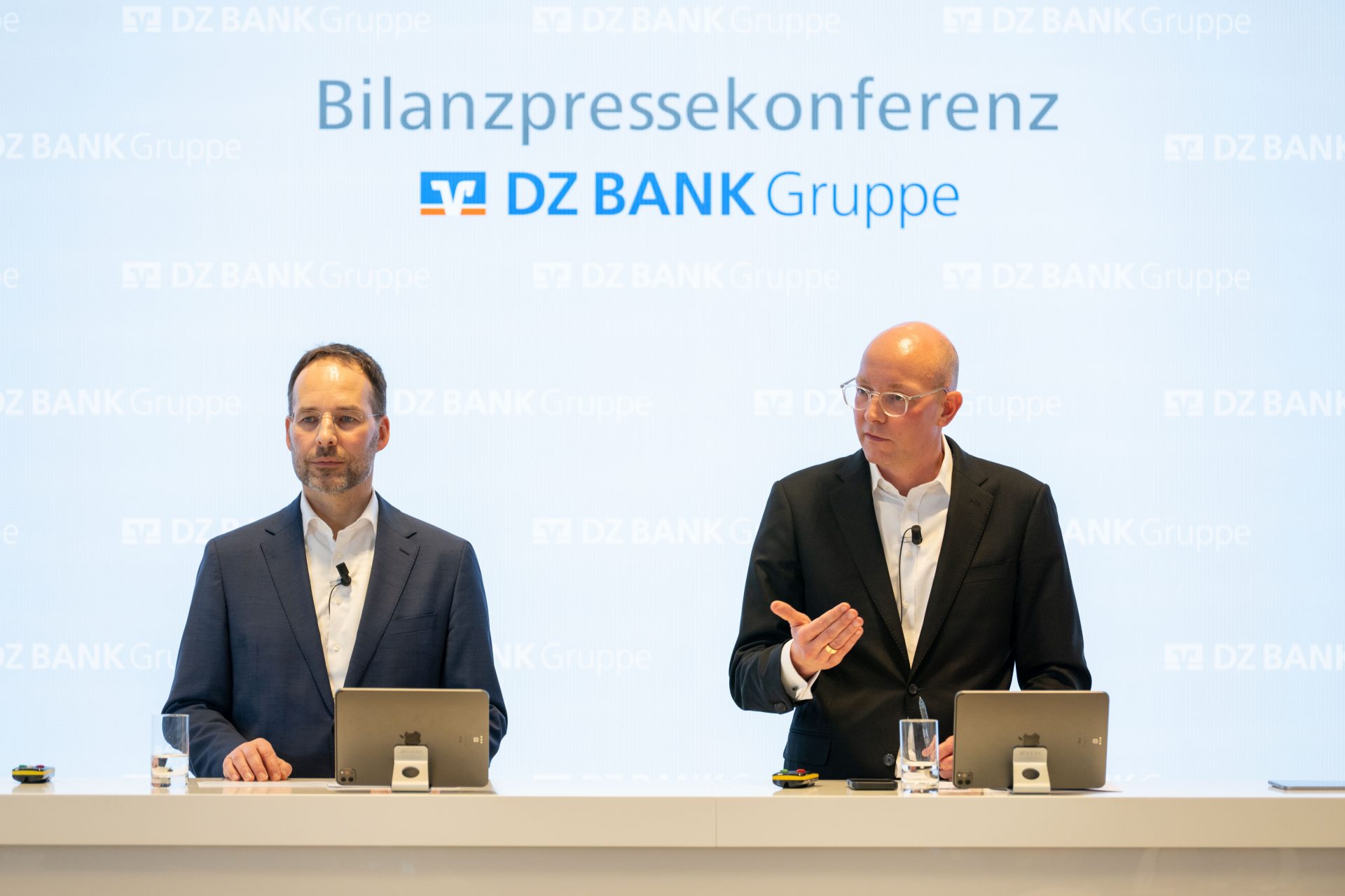 Das Bild zeigt eine Bilanzpressekonferenz der DZ BANK Gruppe. Es sind zwei Personen zu sehen, die an einem Tisch mit Laptops sitzen. Es scheint sich um eine geschäftliche Veranstaltung zu handeln, bei der über finanzielle Ergebnisse berichtet wird.
