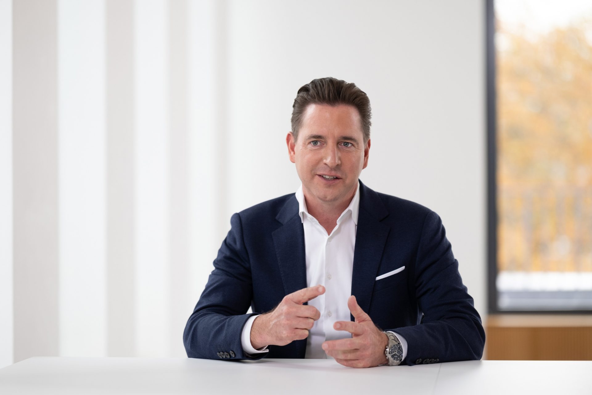 Eine Person in einem dunklen Anzug und weißem Hemd sitzt an einem Tisch und gestikuliert. Der Hintergrund ist hell und enthält ein Fenster mit Blick auf einige Bäume.