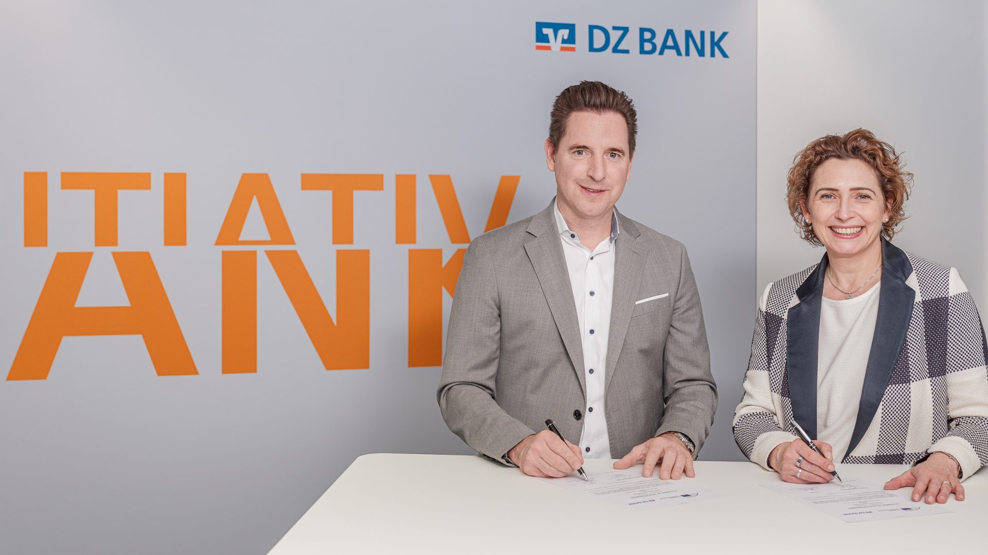 Das Bild zeigt zwei Personen, einen Mann und eine Frau, die nebeneinander an einem weißen Tisch stehen und lächeln. Sie haben beide Dokumente vor sich und halten Stifte in der Hand, was darauf hindeutet, dass sie möglicherweise dabei sind, etwas zu unterzeichnen. Im Hintergrund ist das Logo der DZ BANK zu sehen, daneben steht in großen orangefarbenen Buchstaben ein (teilweise verdecktes) Wort, vermutlich 'INITIATIVE BANK'.