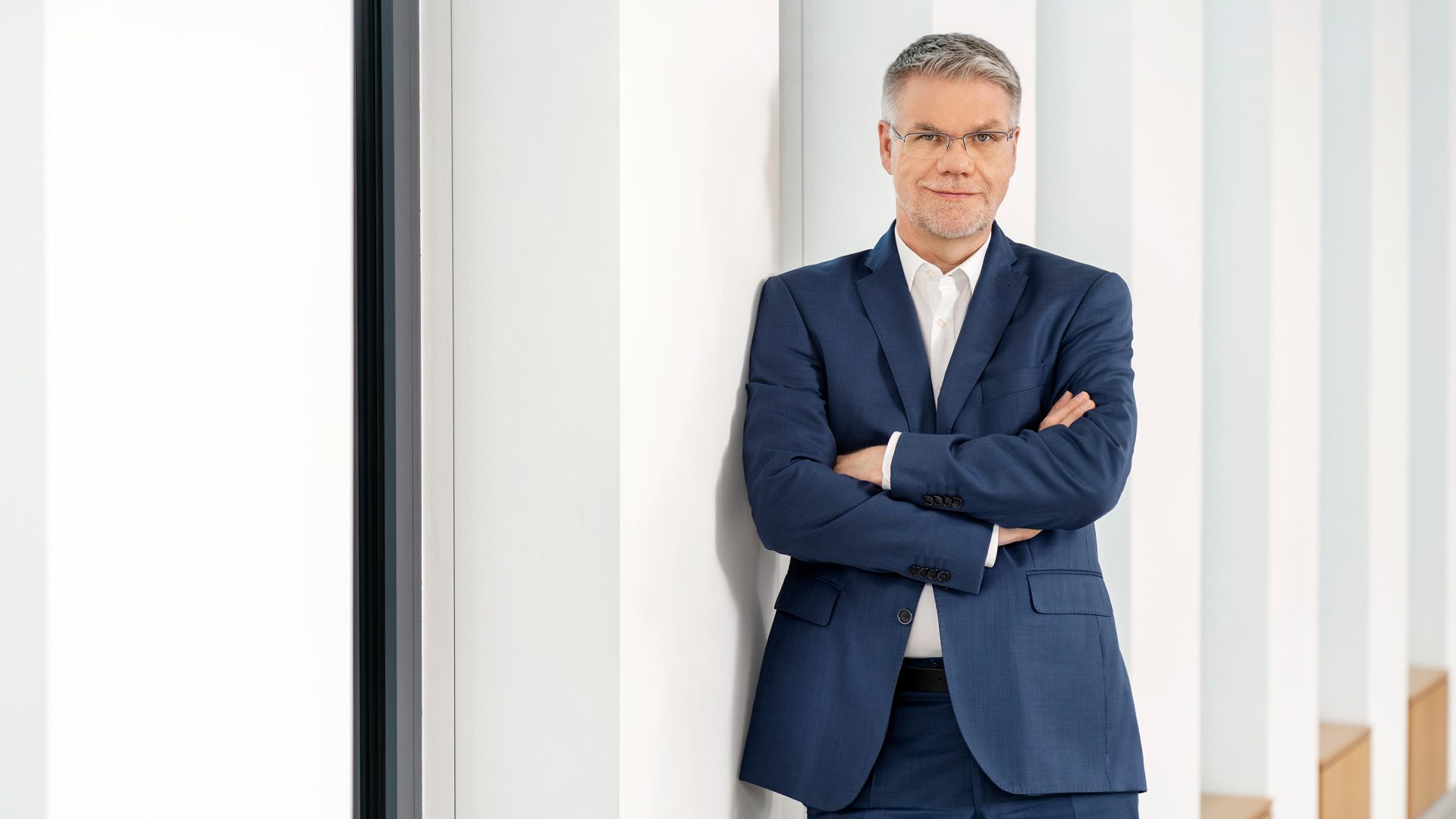 Ein Mann in einem formellen Anzug, der mit verschränkten Armen an einer Wand lehnt, in einem modernen, hellen Raum mit vertikalen weißen Streifen im Hintergrund.