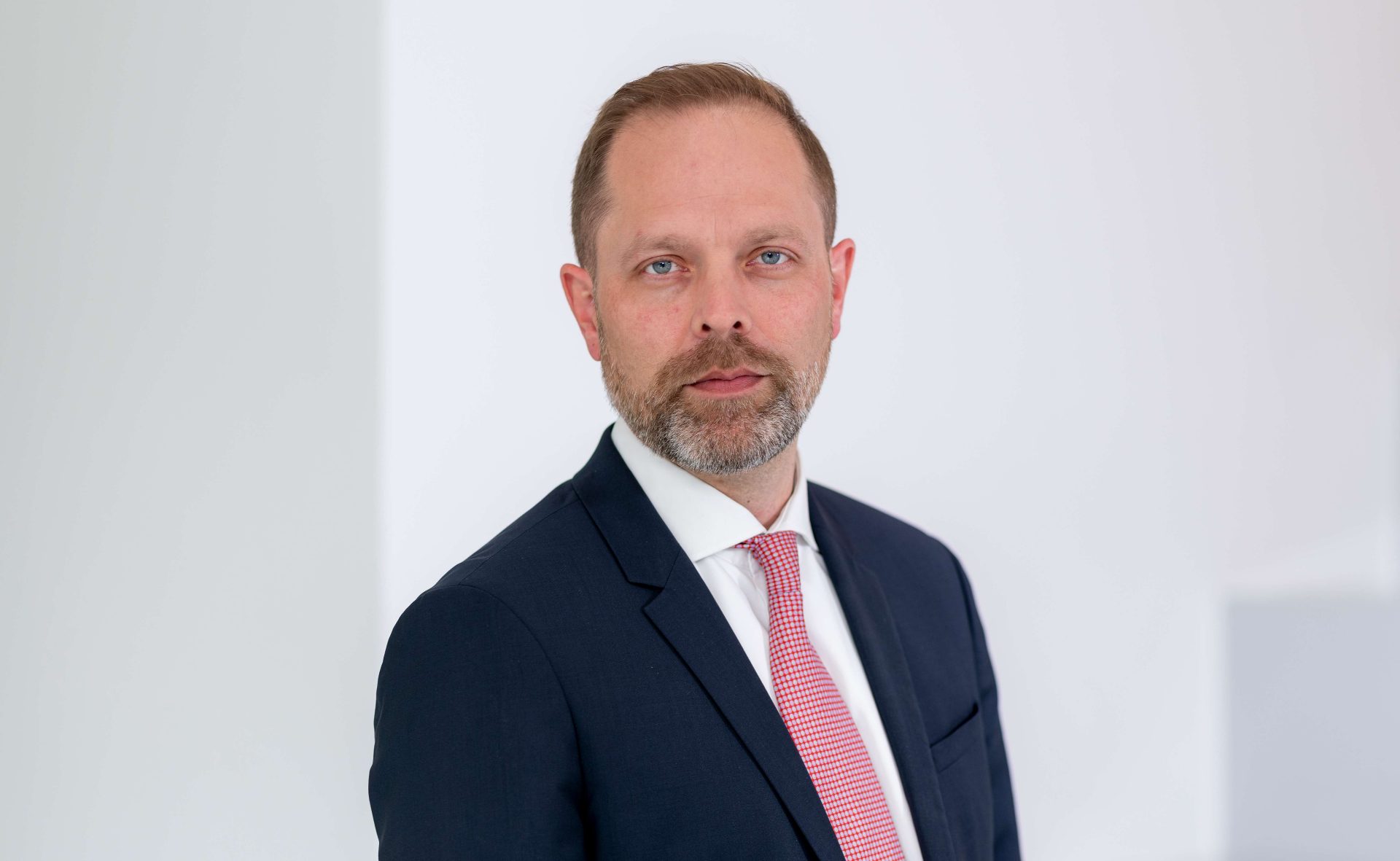 Ein Mann in einem formellen Anzug mit einem weißen Hemd und einer roten Krawatte vor einem weißen Hintergrund.