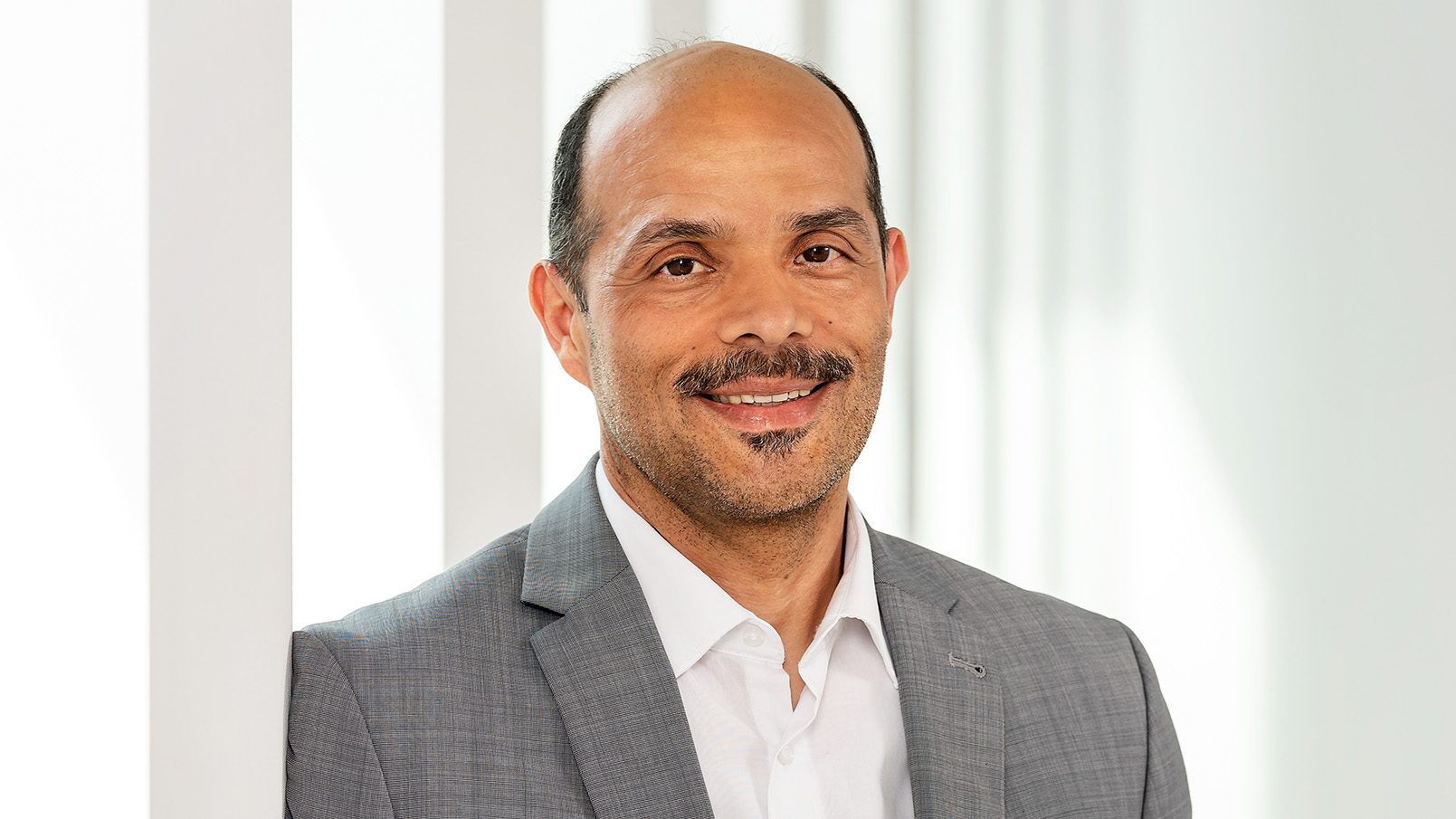 Ein Mann trägt ein formelles graues Sakko über einem weißen Hemd. Er steht vor einem Hintergrund mit vertikalen weißen Linien und scheint in einem hellen Raum zu stehen, möglicherweise ein Büro oder ein professionelles Umfeld.