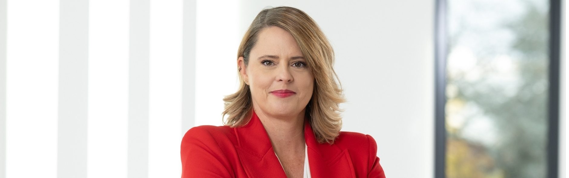 Die Person trägt ein rotes Jackett und befindet sich in einem hellen Raum mit einem Fenster im Hintergrund. Es scheint sich um ein professionelles Porträt oder ein Geschäftsbild zu handeln.