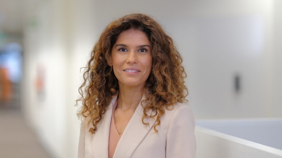 Die Person trägt ein formelles Outfit, bestehend aus einem hellen Blazer und einem Oberteil, und steht anscheinend in einem modernen Bürogebäude oder Korridor mit hellen Wänden und einer klaren Beleuchtung. Es scheint ein professionelles Umfeld zu sein.