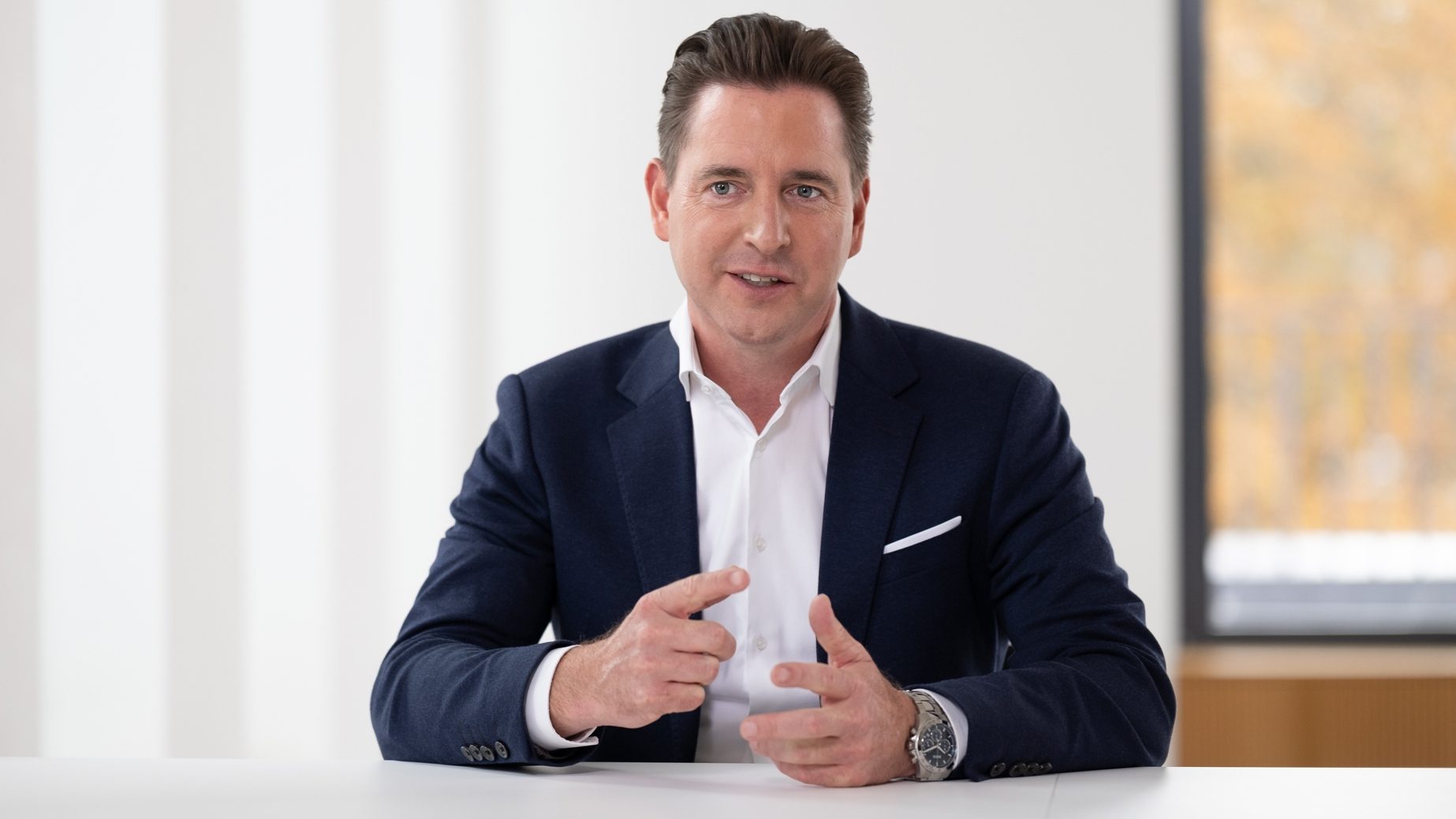 Ein Mann in einem formellen Anzug sitzt an einem Tisch in einem hellen Raum mit einem Fenster, durch das man Herbstlaub sehen kann.