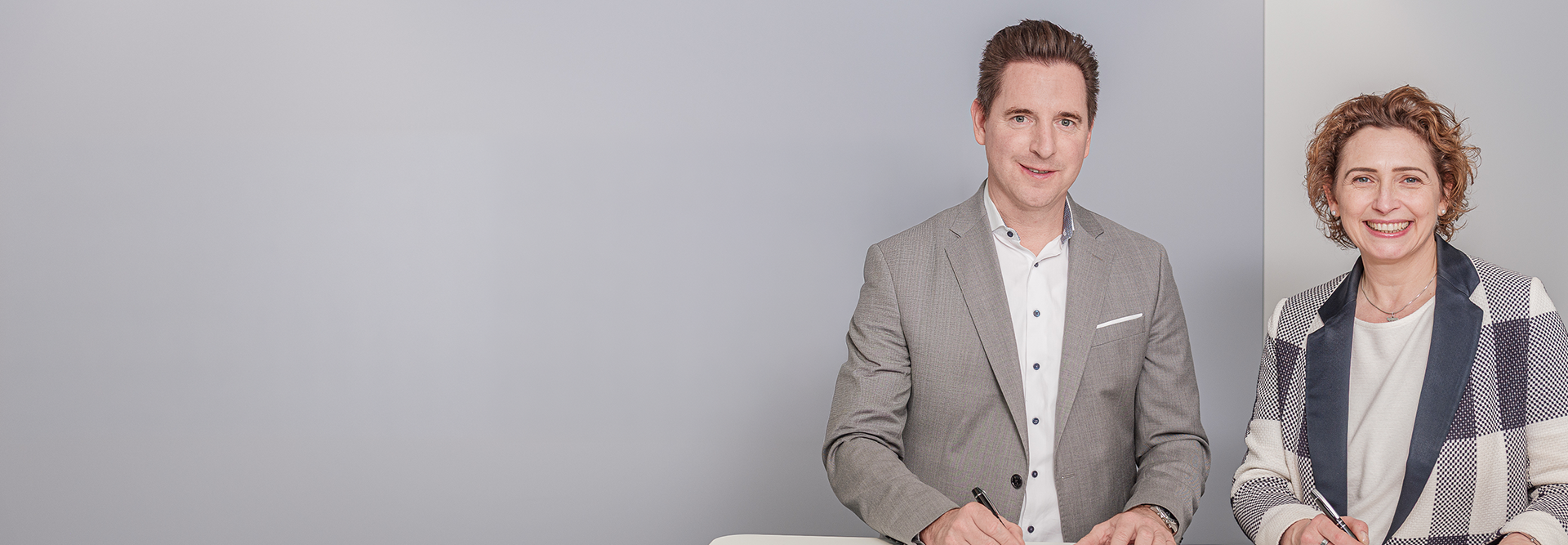 Das Bild zeigt zwei Personen, einen Mann und eine Frau, die nebeneinander sitzen oder stehen und in die Kamera lächeln. Beide sind formell bis business-casual gekleidet. Der Hintergrund ist neutral und hell, was auf einen professionellen Kontext wie ein Büro oder ein Fotoshooting für geschäftliche Zwecke hindeutet.