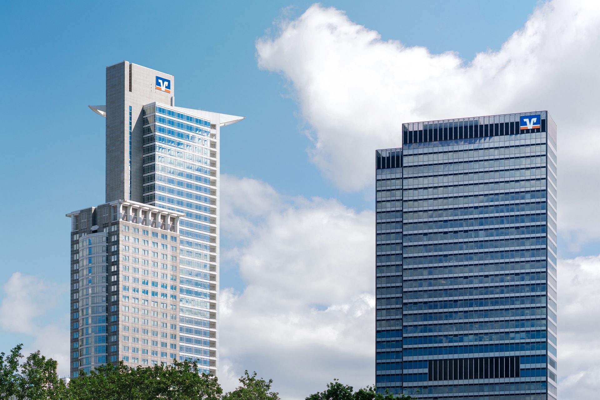Das Bild zeigt zwei moderne Hochhäuser, die wahrscheinlich Teil eines Finanz- oder Geschäftszentrums sind. Beide Gebäude wirken imposant und filigran und sind vor einem klaren Himmel mit einigen Wolken abgebildet. Auf beiden Gebäuden ist das Logo eines Unternehmens zu sehen, das belegt, dass es sich um ein Bankgebäude der Volksbank oder ein ähnliches Finanzinstitut handelt. Die Architektur ist zeitgenössisch und die Glasfassade der Gebäude reflektiert das Sonnenlicht, was ihnen einen glänzenden und modernen Eindruck verleiht. Im Vordergrund sind grüne Bäume zu beobachten, die einen Kontrast zu den urbanen Strukturen bieten und der Umgebung einen Hauch von Natur verleihen.