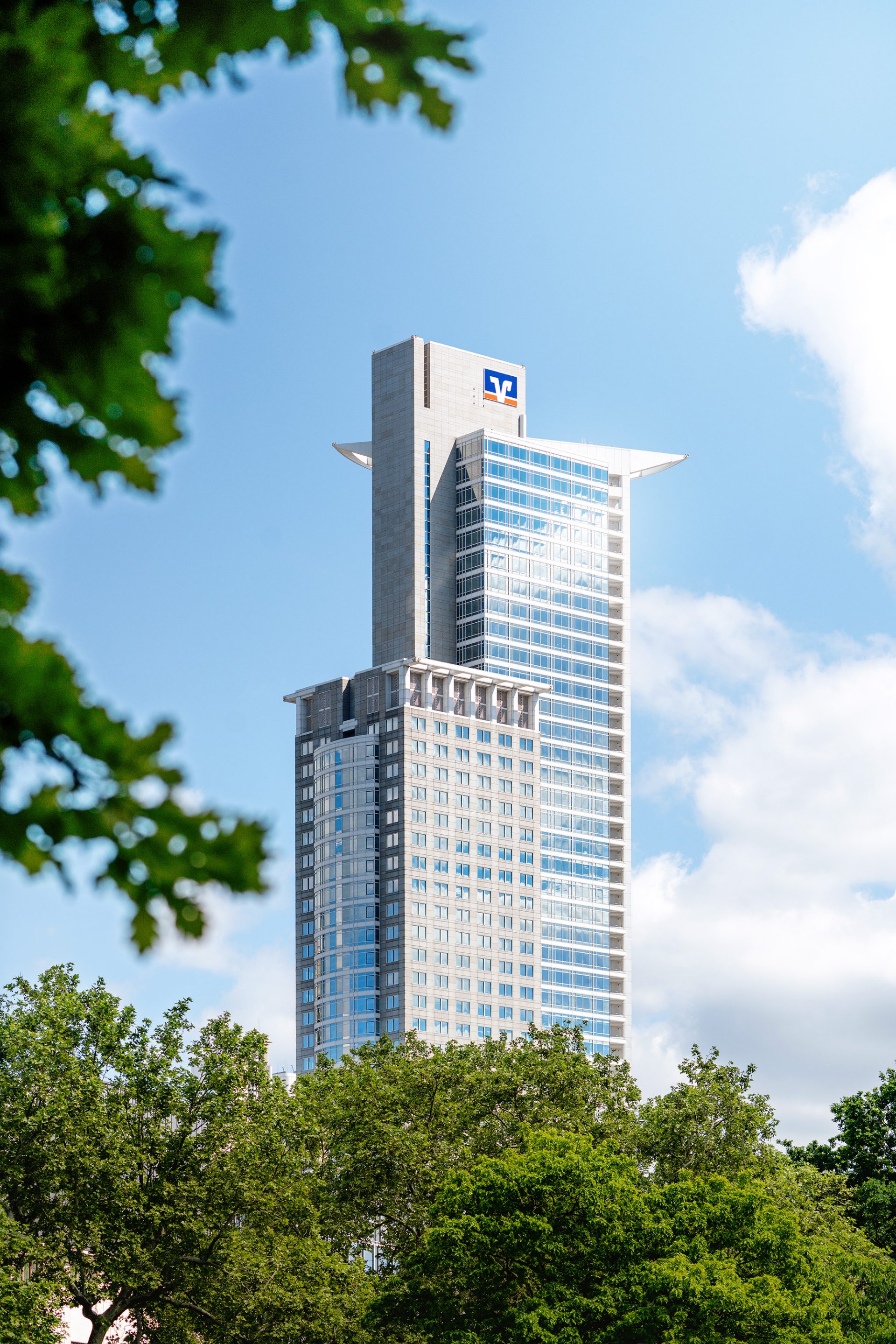 Das Bild zeigt den Victoria-Turm, auch als Marriott-Hotel bekannt, in Frankfurt am Main, Deutschland. Der Turm gehört zur Skyscraper-Gruppe im Stadtteil Innenstadt und ist mit seiner beeindruckenden Architektur und der zentralen Lage eines der Wahrzeichen der Stadt. Die grüne vegetative Umgebung um den Turm herum bildet einen Kontrast zu der modernen städtischen Struktur des Gebäudes.