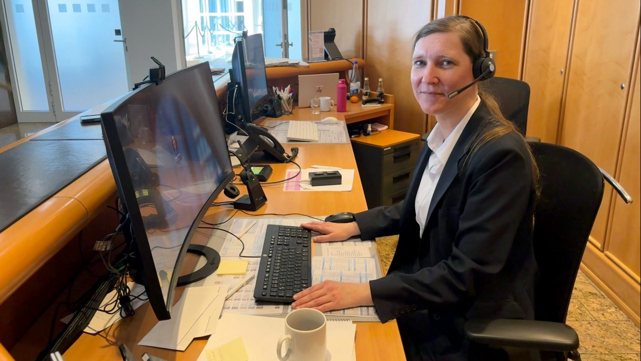 Das Bild zeigt eine Person, die an einem Schreibtisch sitzt und einen Computer mit zwei Monitoren bedient. Sie trägt ein Headset, was darauf hinweist, dass sie möglicherweise telefoniert oder an Videoanrufen teilnimmt. Auf dem Schreibtisch befinden sich Tastatur, Maus, ein Telefon, eine Kaffeetasse und diverse Unterlagen. Der Raum sieht wie ein Büro oder Empfangsbereich aus.