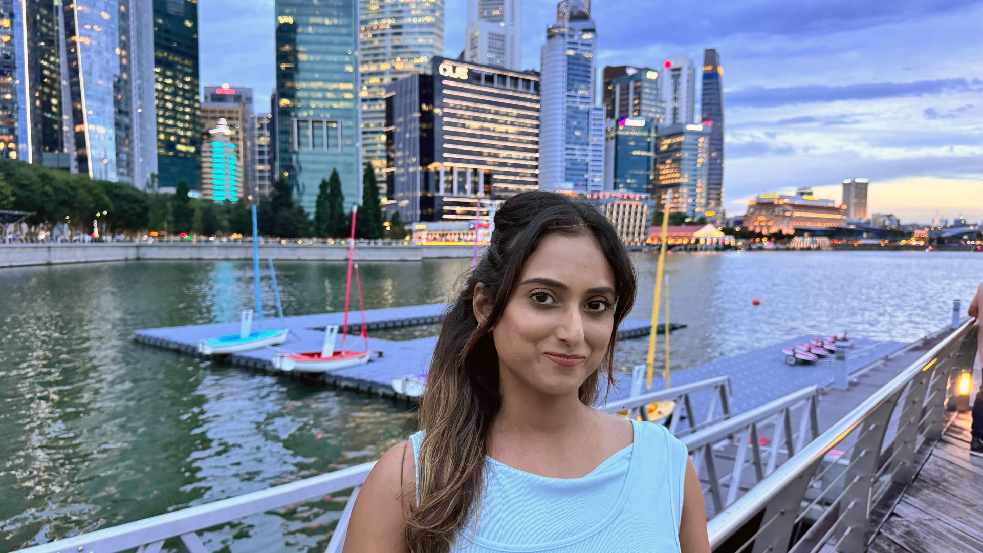 Das Bild zeigt eine städtische Umgebung am Wasser mit modernen Hochhäusern und einem Steg, auf dem Boote angedockt sind. Es ist wahrscheinlich der Marina Bay in Singapur.