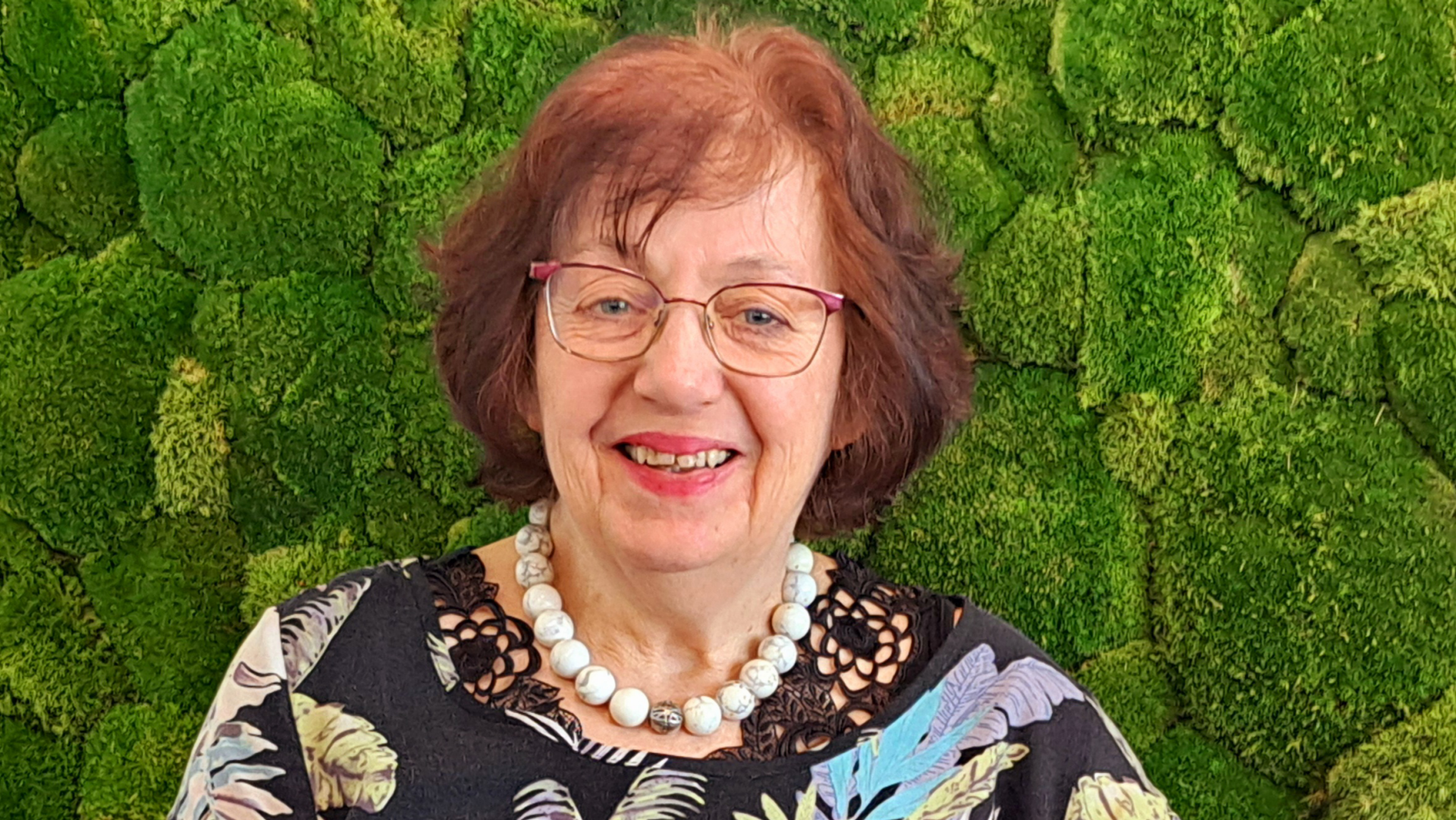 Die Aufnahme zeigt eine Person vor einem hellgrünen Hintergrund aus Moos oder ähnlich strukturiertem Material. Die Person trägt eine Halskette aus großen weißen Perlen und ein Kleidungsstück mit Blumenmuster, das in verschiedenen Farben gehalten ist.