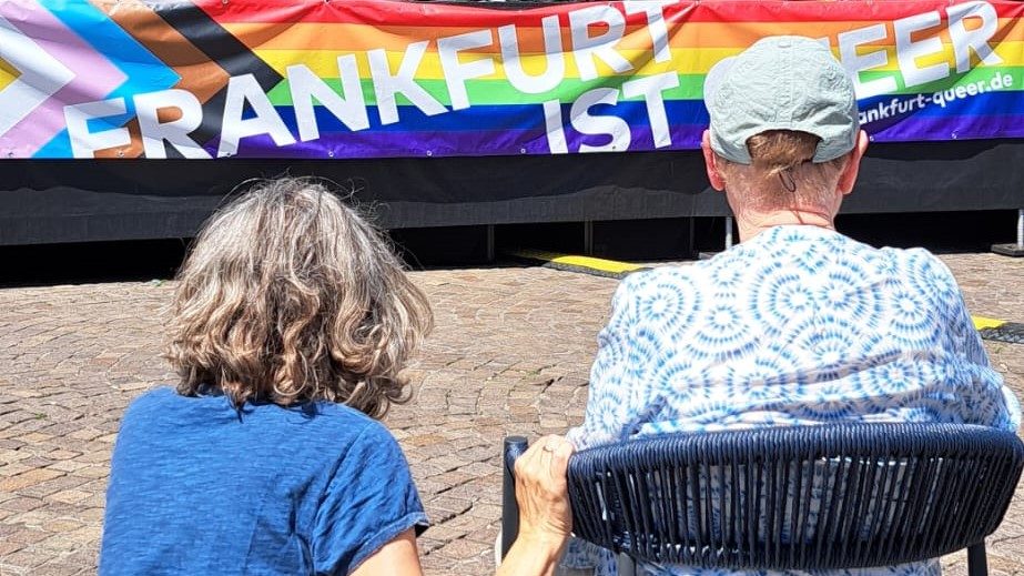 Das Bild zeigt zwei Personen mit dem Rücken zur Kamera, scheinbar auf Stühlen oder Bänken sitzend, vor einem bunten Banner. Das Banner trägt die Aufschrift "FRANKFURT IST QUEER" und ist in den Farben der Regenbogenflagge gestaltet, die ein Symbol für die LGBTQ+ Gemeinschaft ist. 

**Beschreibung der Bilddetails:**
- **Personen:** Zwei Menschen, von hinten gesehen, ruhig sitzend.
- **Ort:** Eine gepflasterte Fläche im Freien, wahrscheinlich eine Veranstaltung oder ein öffentlicher Platz.
- **Banner:** Groß und über dem Boden, auffällig in Regenbogenfarben.
- **Text auf dem Banner:** "FRANKFURT IST QUEER"
- **Symbolik:** Verwendung der Regenbogenfarben deutet auf eine LGBTQ+ Veranstaltung oder Feier hin.
  
**Interpretation:**
Das Bild könnte eine Feier, Parade oder ein Event in Frankfurt am Main darstellen, das die Vielfalt und das LGBTQ+ Leben feiert. Der einfache und kraftvolle Text unterstreicht ein stolzes Statement für die Stadt als Ort der Akzeptanz und Inklusivität.