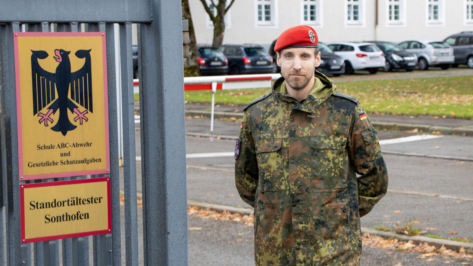 Ein Soldat der Bundeswehr steht vor einem Schild, das den Standortältesten von Sonthofen und die Schule ABC-Ausb und GrundlFhr beheimatet kennzeichnet. Das Bild zeigt den Eingangsbereich eines militärischen Standortes in Deutschland mit geparkten Autos im Hintergrund.