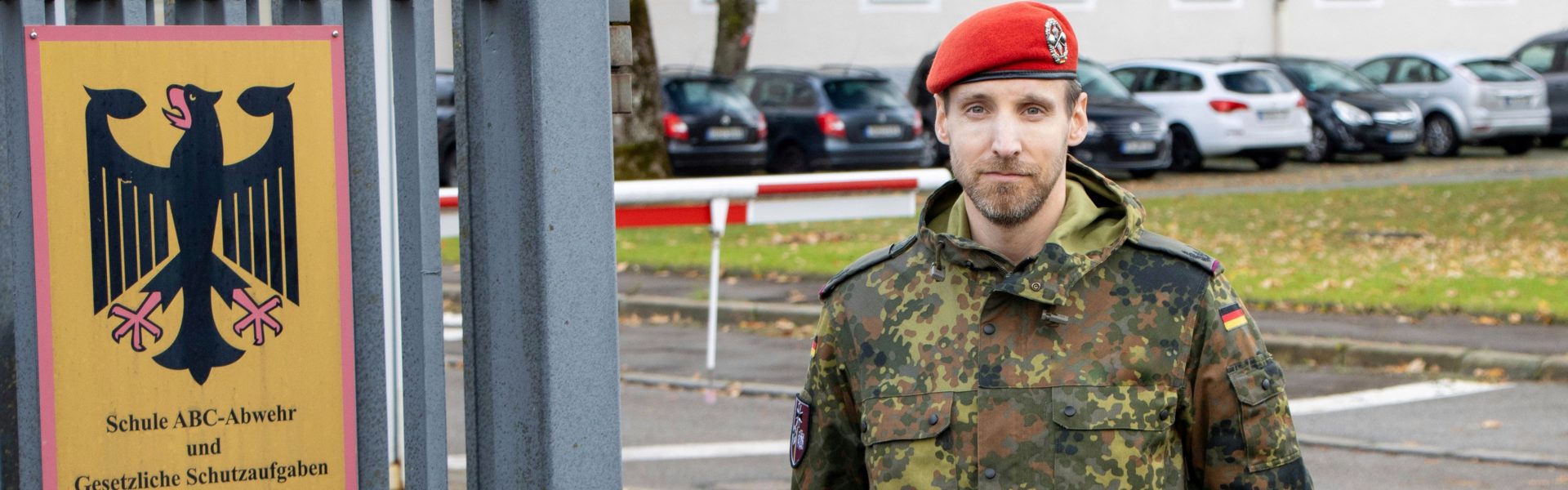 Ein Soldat der Bundeswehr in einer militärischen Uniform mit einer roten Kopfbedeckung, vor einem Schild mit einem Bundesadler und dem Schriftzug "Schule ABC-Abwehr und Gesetzliche Schutzaufgaben". Hintergrund zeigt geparkte Autos und eine Schranke.