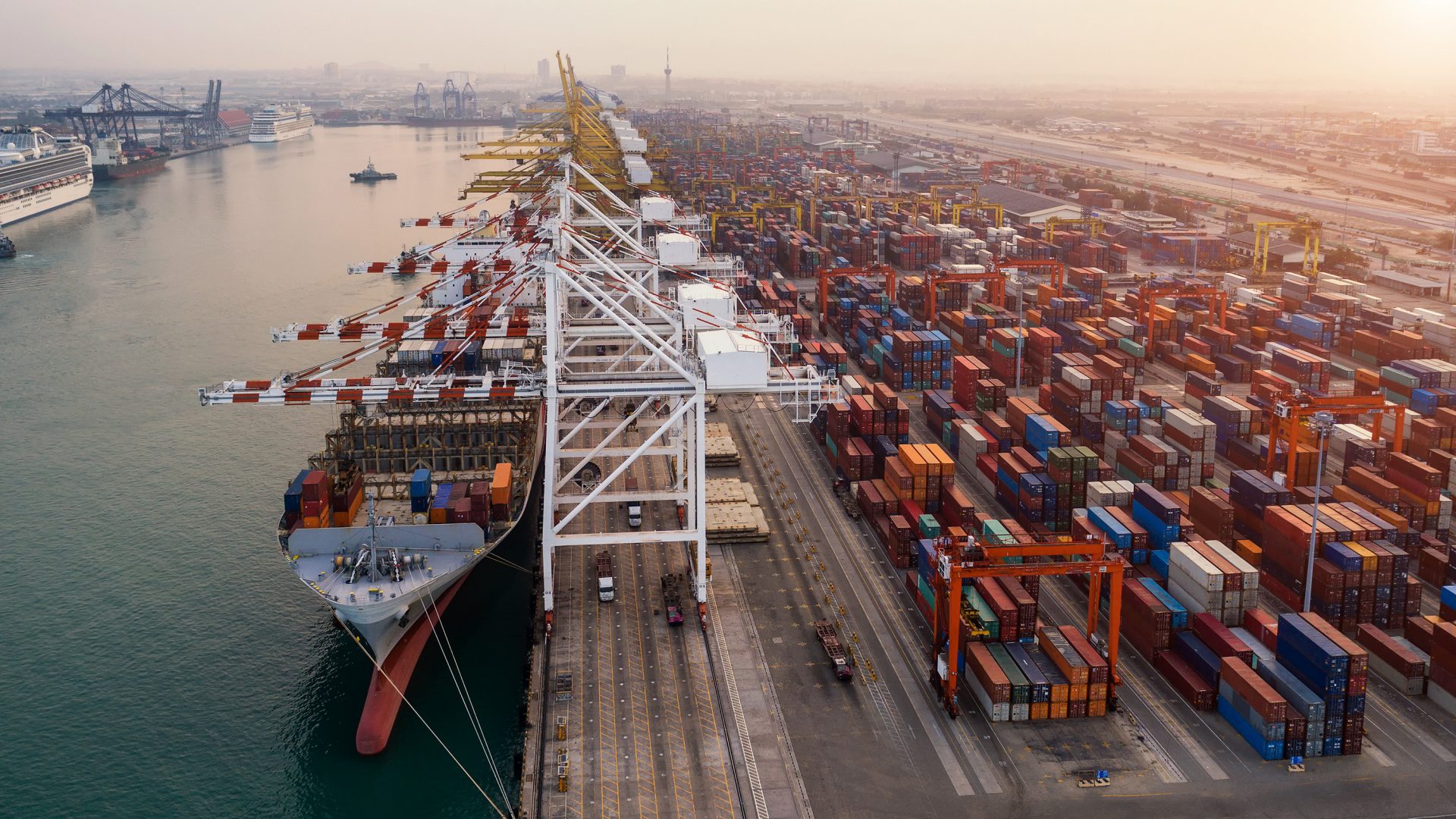 Das Bild zeigt einen großen Containerhafen. Dort sind zahlreiche Container zu sehen, sowie Kräne, die für das Be- und Entladen von Containerschiffen verwendet werden. Ein großes Containerschiff liegt am Kai und wird vermutlich gerade be- oder entladen. Im Hintergrund sind weitere Hafenanlagen und zahlreiche Container zu erkennen.