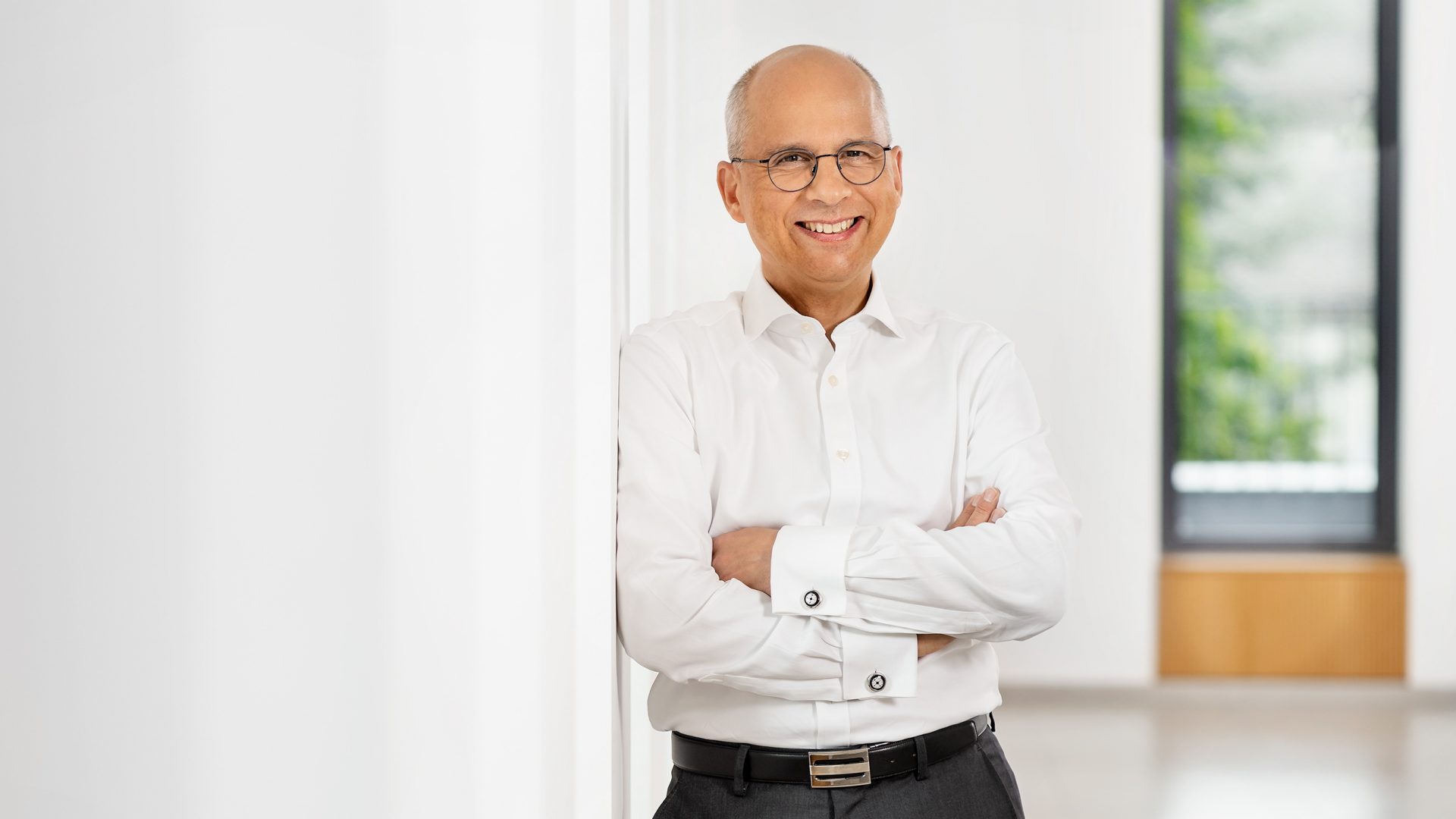 Die Aufnahme zeigt eine Person in einem hellen Raum, die mit verschränkten Armen links neben einem großen Fenster steht. Die Person trägt ein weißes Hemd und dunkle Hosen. Der Hintergrund ist teilweise unscharf, mit einem Fenster und einigen grünen Pflanzen außerhalb des Fensters sichtbar. Das Bild vermittelt eine berufliche Atmosphäre.