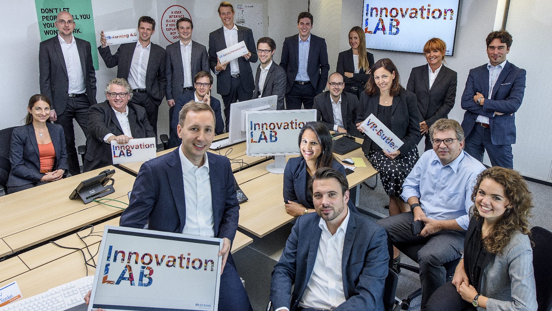 Das Bild zeigt eine Gruppe von Menschen in einem modernen Büro oder Konferenzraum, die gemeinsam posieren. Mehrere Personen halten Schilder mit der Aufschrift 'Innovation LAB' in den Händen, und auf Monitoren im Hintergrund steht ebenfalls 'Innovation LAB'. Es handelt sich offenbar um ein Team oder eine Gruppe, die an einem Innovationsprojekt arbeitet oder an einer Innovationsveranstaltung teilnimmt.