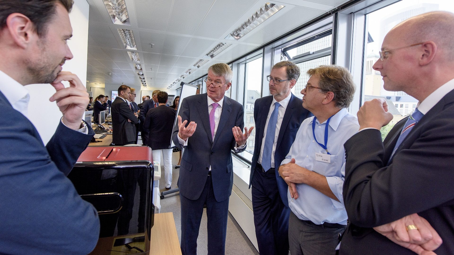Das Bild zeigt eine kleine Gruppe von Männern in formeller Kleidung, die sich in einem modernen Büro- oder Konferenzraum unterhalten. Im Hintergrund sind weitere Personen zu sehen, die ebenfalls miteinander sprechen. Die Atmosphäre wirkt professionell, möglicherweise handelt es sich um ein geschäftliches Treffen, eine Konferenz oder eine Fachveranstaltung.