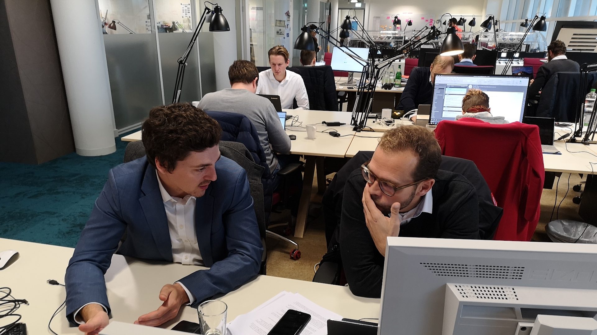 Das Bild zeigt ein modernes Großraumbüro, in dem mehrere Personen an Schreibtischen arbeiten. Im Vordergrund unterhalten sich zwei Männer, die vor Computermonitoren sitzen. Im Hintergrund arbeiten weitere Personen an ihren Arbeitsplätzen, viele Computer und Büromaterialien sind zu sehen. Das Gesamtbild vermittelt eine geschäftige Arbeitsatmosphäre.