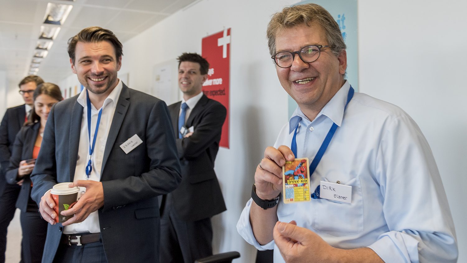 Das Bild zeigt eine Gruppe von Menschen in Business-Kleidung in einem Büro oder Konferenzraum. Zwei Männer stehen im Vordergrund, einer hält einen Kaffee-Becher und lacht, der andere zeigt eine bunte Karte und lächelt ebenfalls. Im Hintergrund stehen weitere Personen, es herrscht eine freundliche und entspannte Atmosphäre. Es scheint sich um eine geschäftliche oder berufliche Veranstaltung oder ein Treffen zu handeln.
