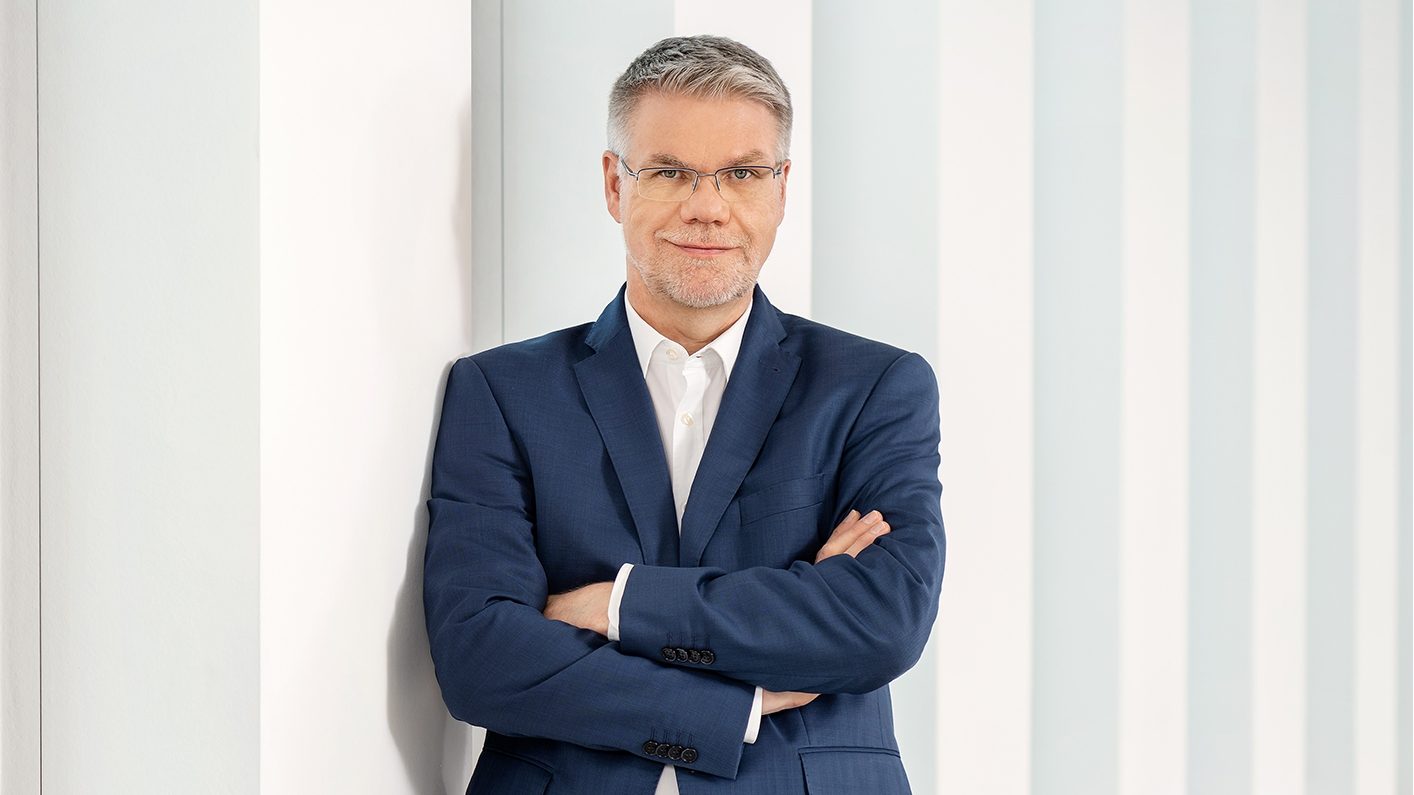 Die Aufnahme zeigt eine Person in einem hellblauen Anzug mit verschränkten Armen, die vor einem geometrisch gestreiften Hintergrund steht. Der Anzug vermittelt einen professionellen Eindruck, was möglicherweise auf ein geschäftliches oder formelles Umfeld hinweist.