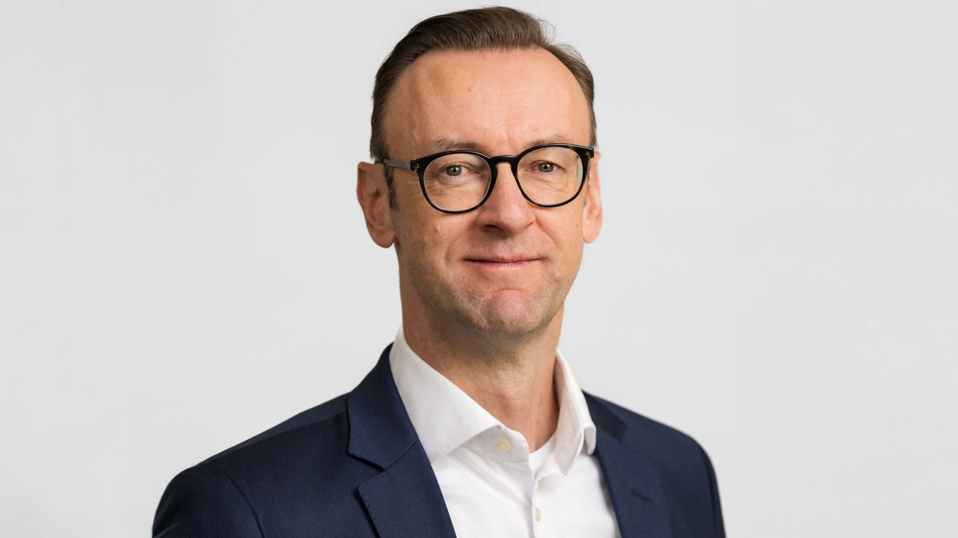 Das Bild zeigt einen Mann mit Brille, der einen dunklen Anzug und ein weißes Hemd trägt. Er steht vor einem neutralen, hellen Hintergrund und schaut in die Kamera. Es handelt sich um ein professionell aufgenommenes Portraitfoto.