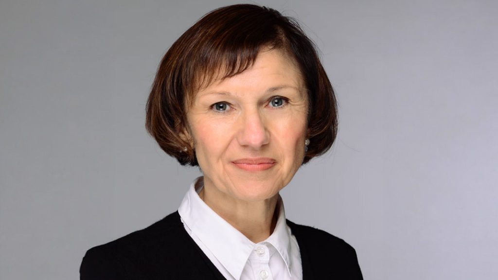 Das Bild zeigt eine Frau im mittleren Alter vor einem neutralen, hellgrauen Hintergrund. Sie trägt einen dunklen Blazer und eine weiße Bluse und schaut freundlich in die Kamera. Das Bild wirkt wie ein professionelles Porträtfoto, möglicherweise für berufliche Zwecke, wie z. B. ein Bewerbungsfoto oder ein Mitarbeiterfoto.