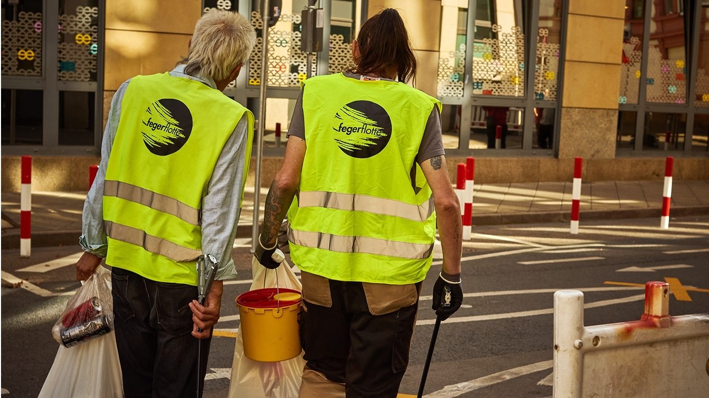 Das Bild zeigt zwei Personen in gelben Warnwesten, die auf einer Straße gehen. Sie tragen Müllsäcke, einen Eimer und Greifwerkzeuge, was darauf hindeutet, dass sie Müll sammeln. Die Szene wirkt wie eine Aktion zur Straßenreinigung oder ein Umweltprojekt in einer städtischen Umgebung.