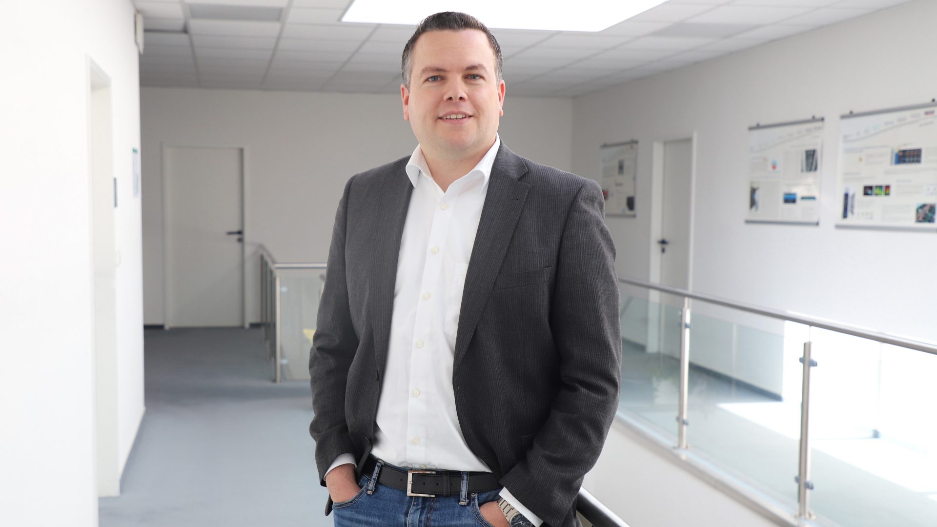 Das Bild zeigt einen Mann, der in einem modernen Bürogebäude oder Flur steht. Er trägt einen dunklen Blazer, ein weißes Hemd und Jeans. Im Hintergrund sind Wände mit Postern oder Informationsmaterialien sowie Türen zu sehen. Die Umgebung wirkt professionell und hell beleuchtet.