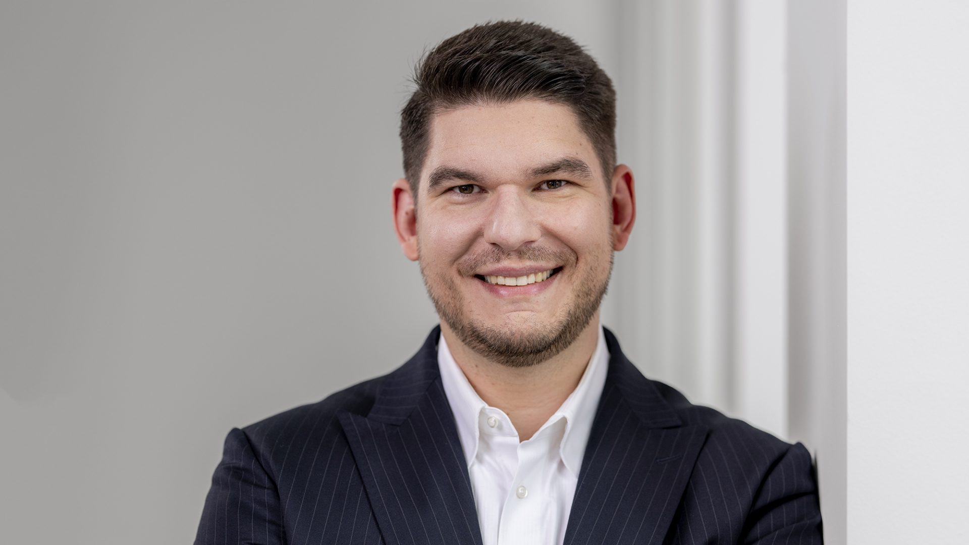 Das Bild zeigt einen Mann in einem dunklen Anzug mit weißem Hemd, der freundlich lächelt. Er steht vor einem neutralen, hellen Hintergrund.