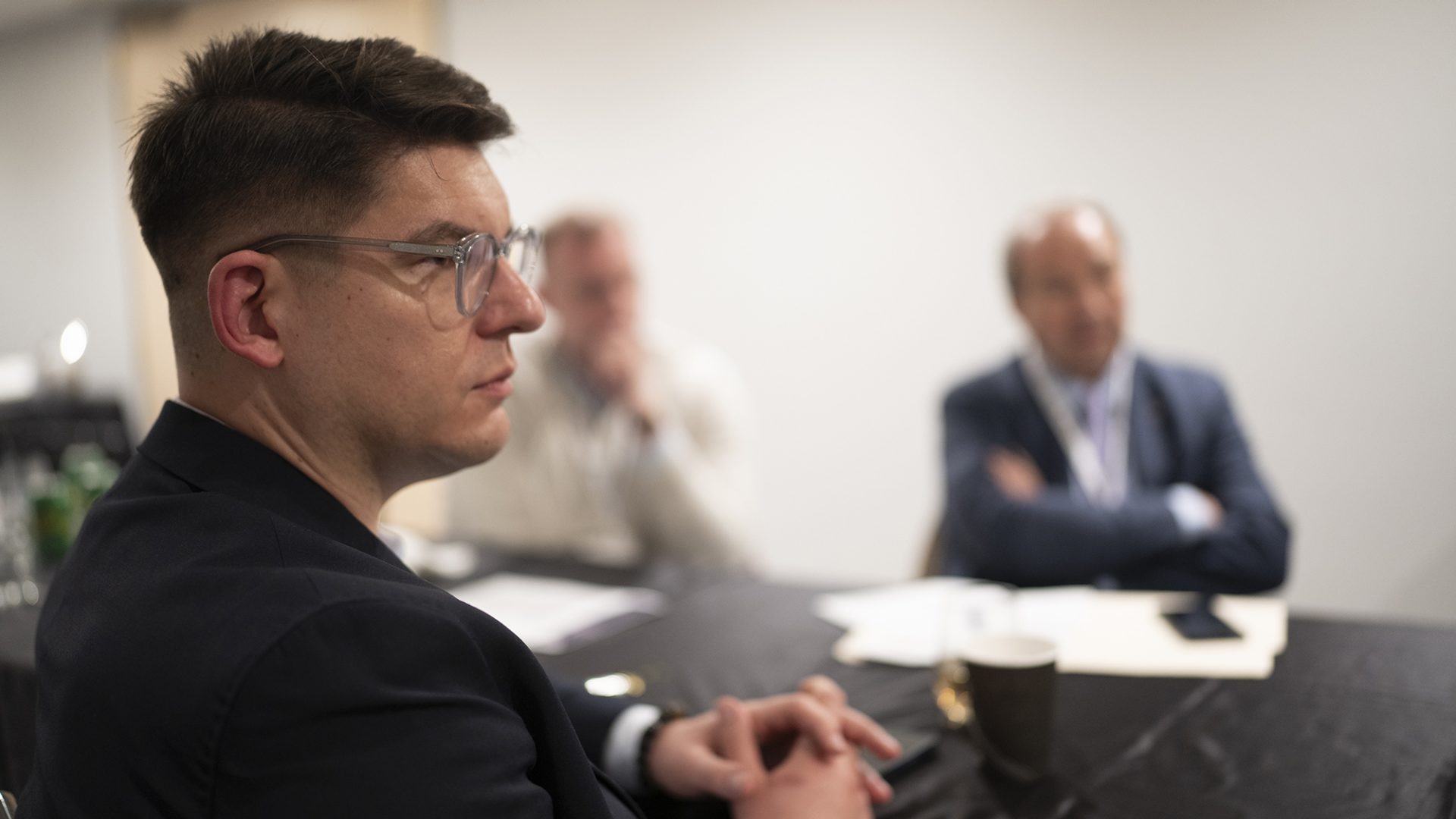 Das Bild zeigt eine Gruppe von Männern, die an einem Tisch sitzen und vermutlich an einem Treffen oder einer Besprechung teilnehmen. Im Vordergrund ist ein Mann mit Brille und dunklem Sakko scharf, während die anderen Personen im Hintergrund unscharf sind. Auf dem Tisch befinden sich Papiere, eine Kaffeetasse und andere Gegenstände.