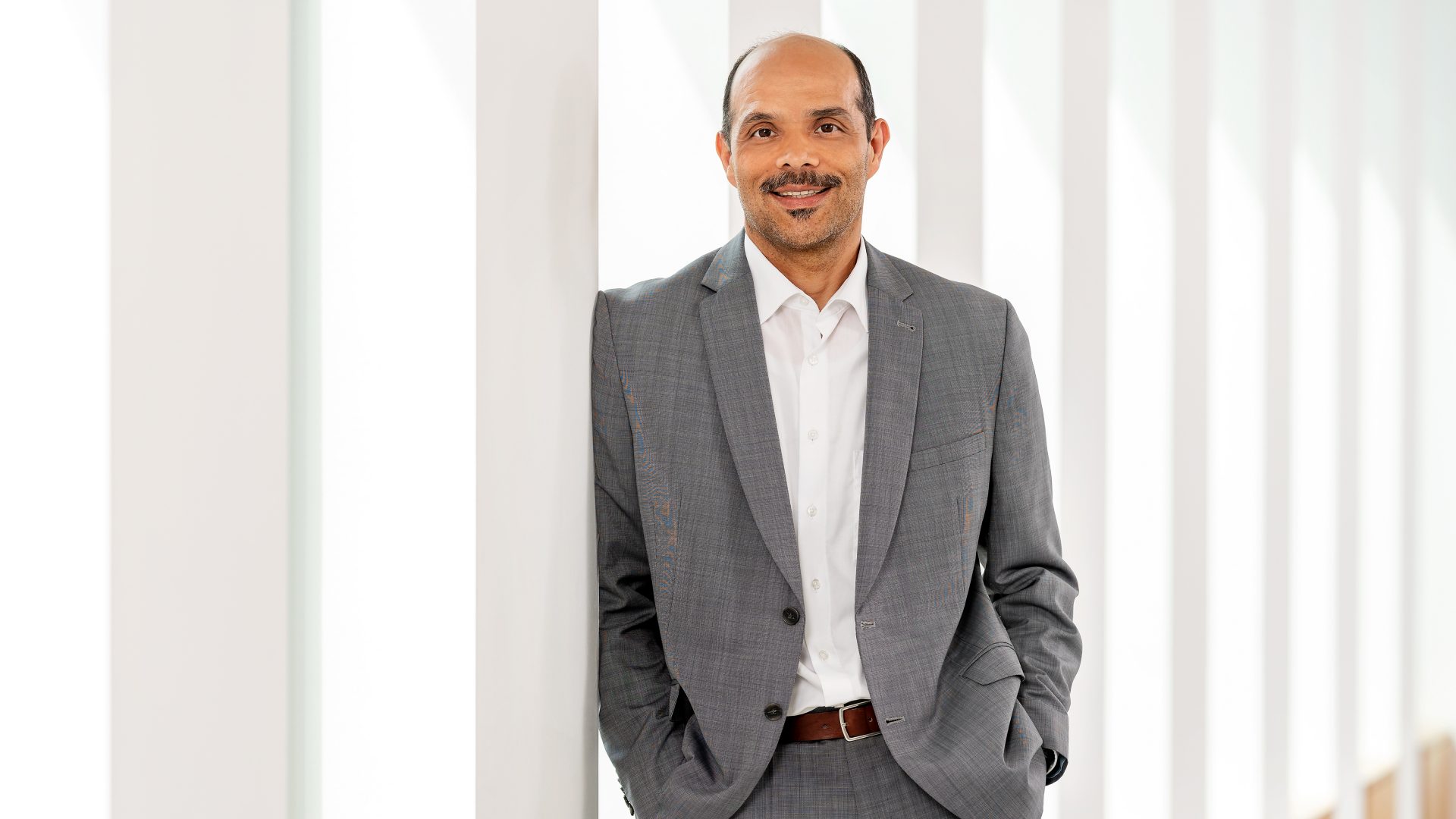 Eine Person, die einen grauen Anzug und ein weißes Hemd trägt. Sie steht in einer Reihe von Säulen oder einem modernen architektonischen Raum.