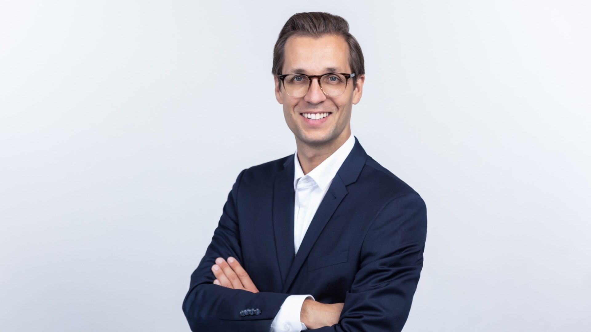 Das Bild zeigt eine Person in einem dunklen Anzug und weißem Hemd vor einem hellen Hintergrund. Die Person steht mit verschränkten Armen und lächelt in die Kamera.