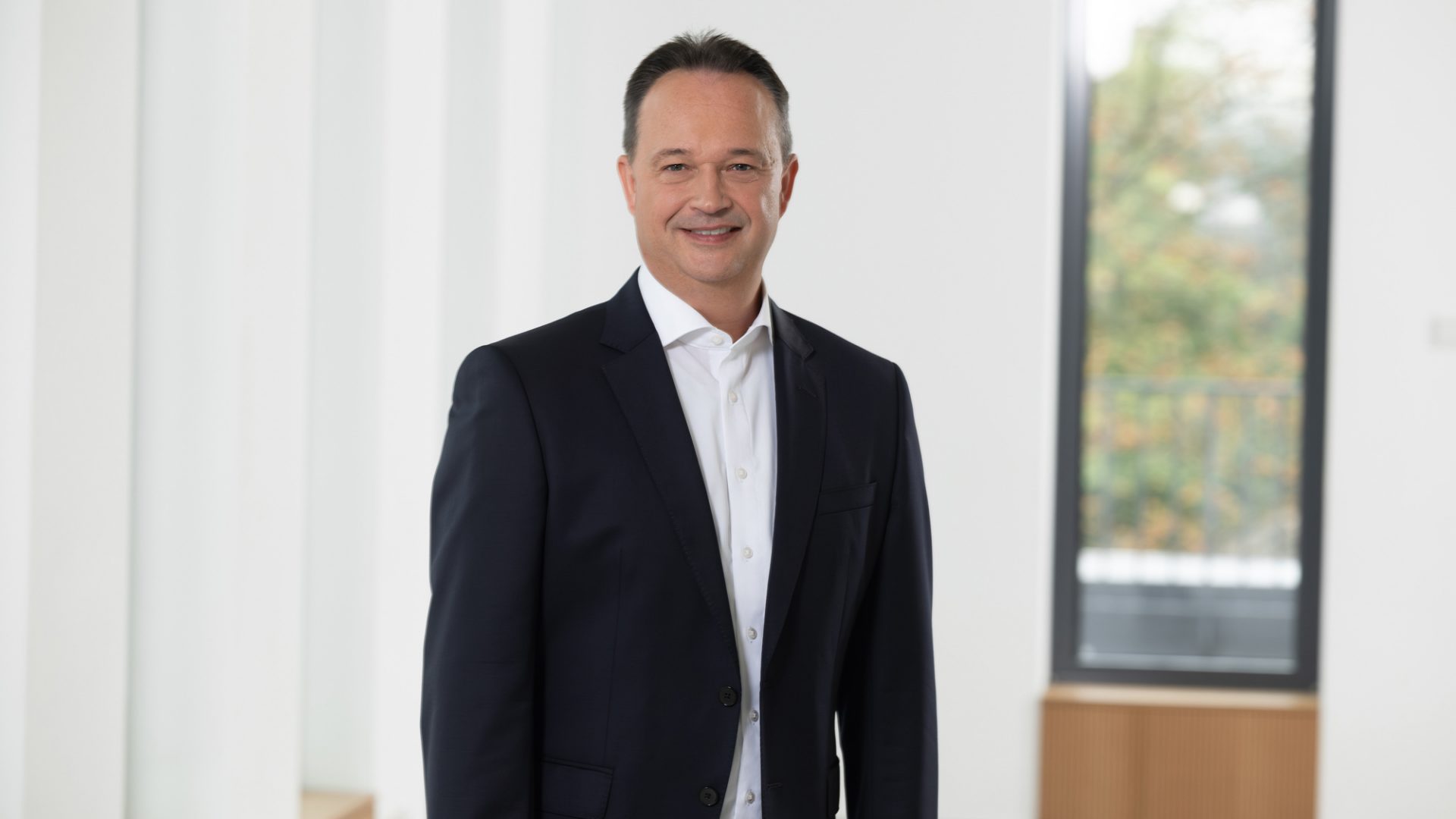 Das Bild zeigt eine Person in einem dunklen Anzug mit weißem Hemd. Der Hintergrund ist in einem hellen Innenbereich mit Fenstern.