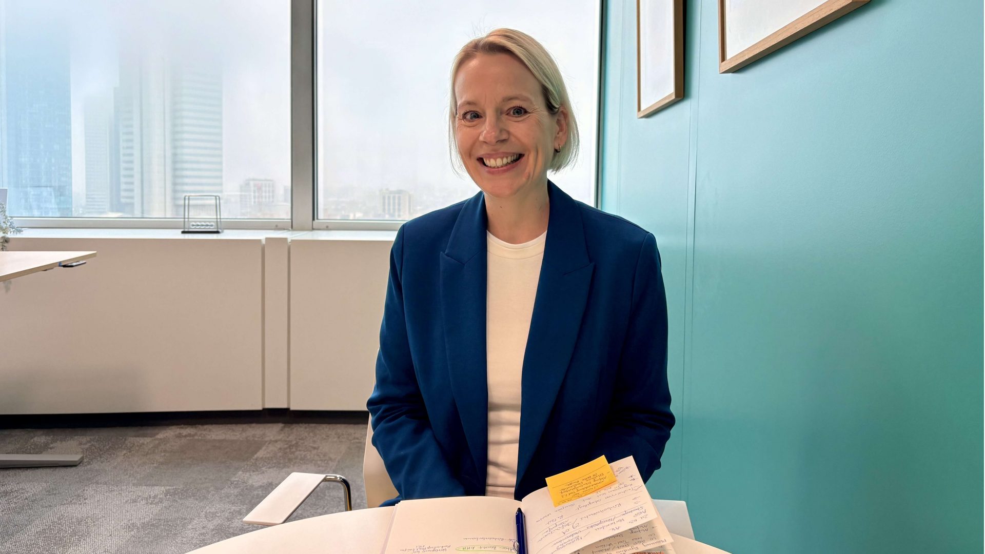 Das Bild zeigt eine Person, die an einem Tisch mit einem aufgeschlagenen Buch und Notizen sitzt. Es gibt Fenster im Hintergrund sowie einige Bilder an der Wand.