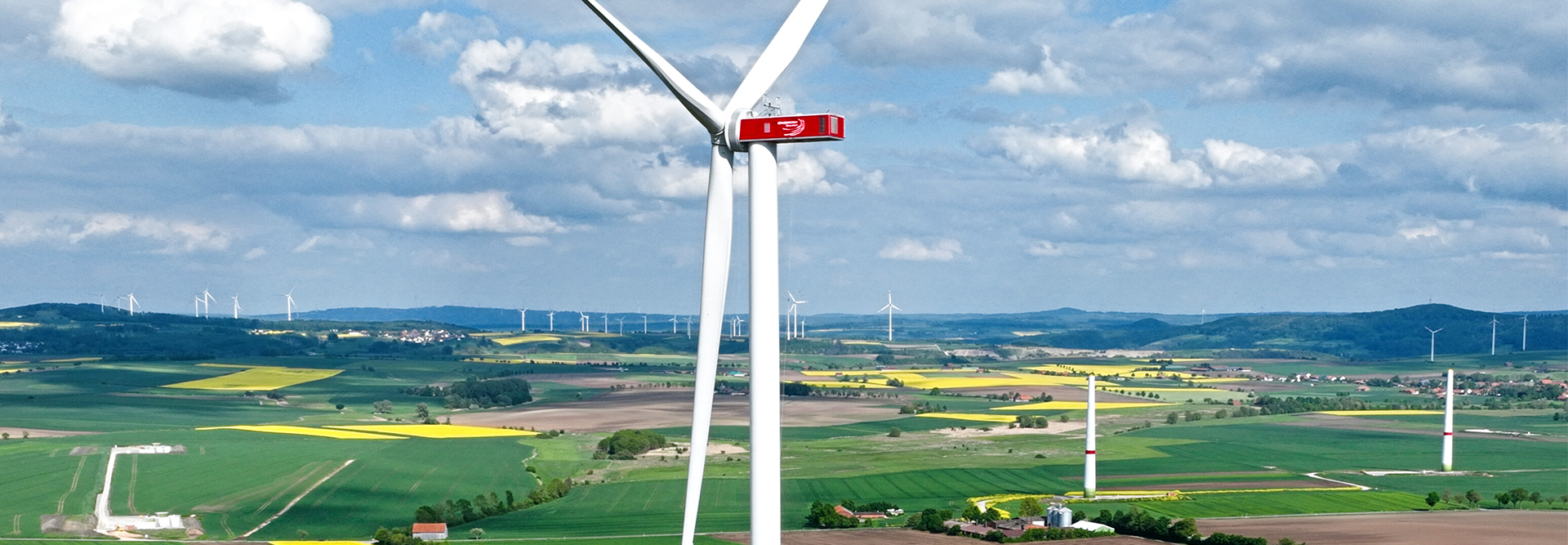 Das Bild zeigt eine Windkraftanlage in einer ländlichen Landschaft mit mehreren anderen Windrädern im Hintergrund. Es gibt Felder und landwirtschaftliche Flächen unter einem blauen Himmel mit einigen Wolken.
