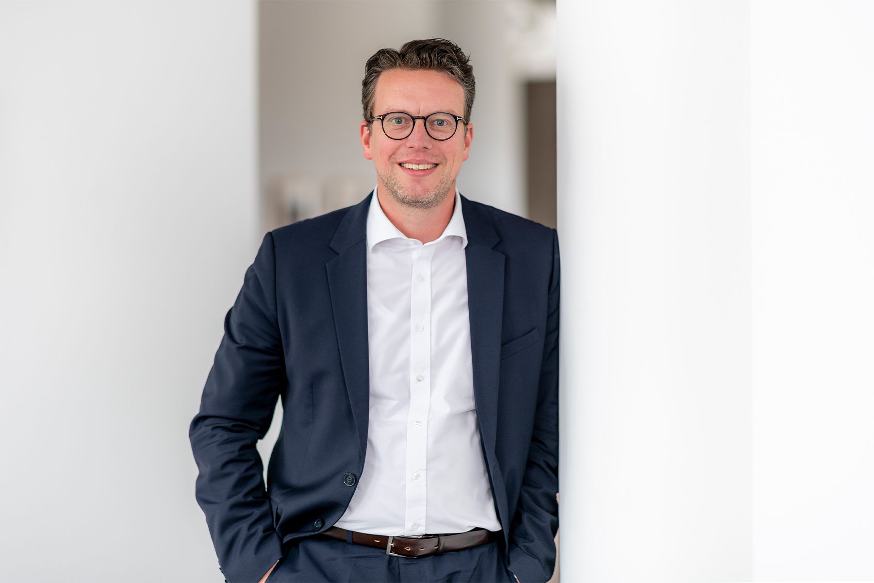 Ein Mann in einem formellen Anzug, der an eine weiße Wand lehnt. Er trägt ein dunkelblaues Sakko und eine dazu passende Hose, kombiniert mit einem weißen Hemd und einem braunen Ledergürtel. Der Raum scheint modern und hell beleuchtet zu sein.