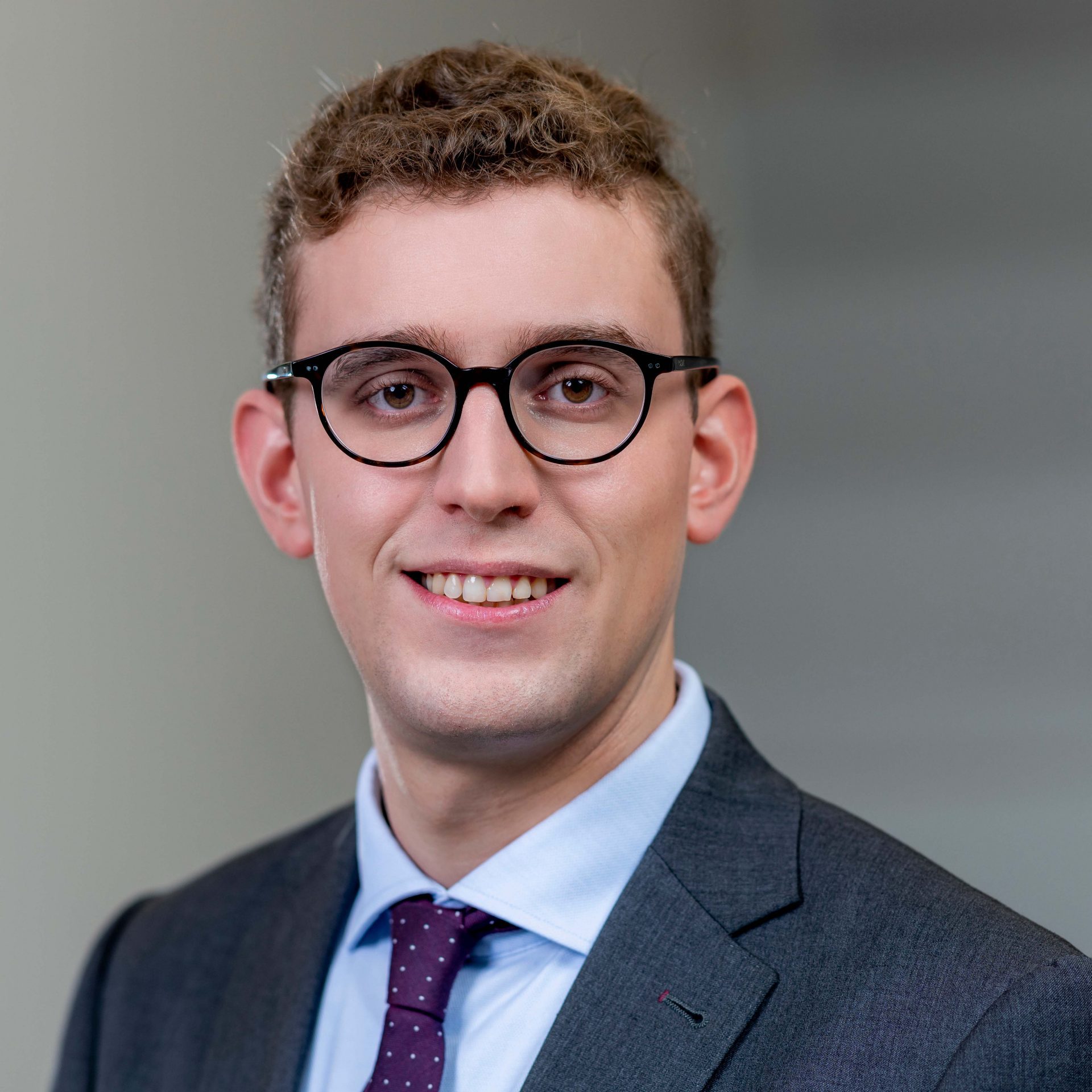 Das Bild zeigt einen jungen Mann mit lockigen Haaren und Brille, der einen Anzug, ein Hemd und eine Krawatte trägt. Er steht vor einem neutralen Hintergrund und lächelt in die Kamera. Es handelt sich vermutlich um ein professionelles Porträtfoto, wie es in geschäftlichen oder akademischen Kontexten verwendet wird.