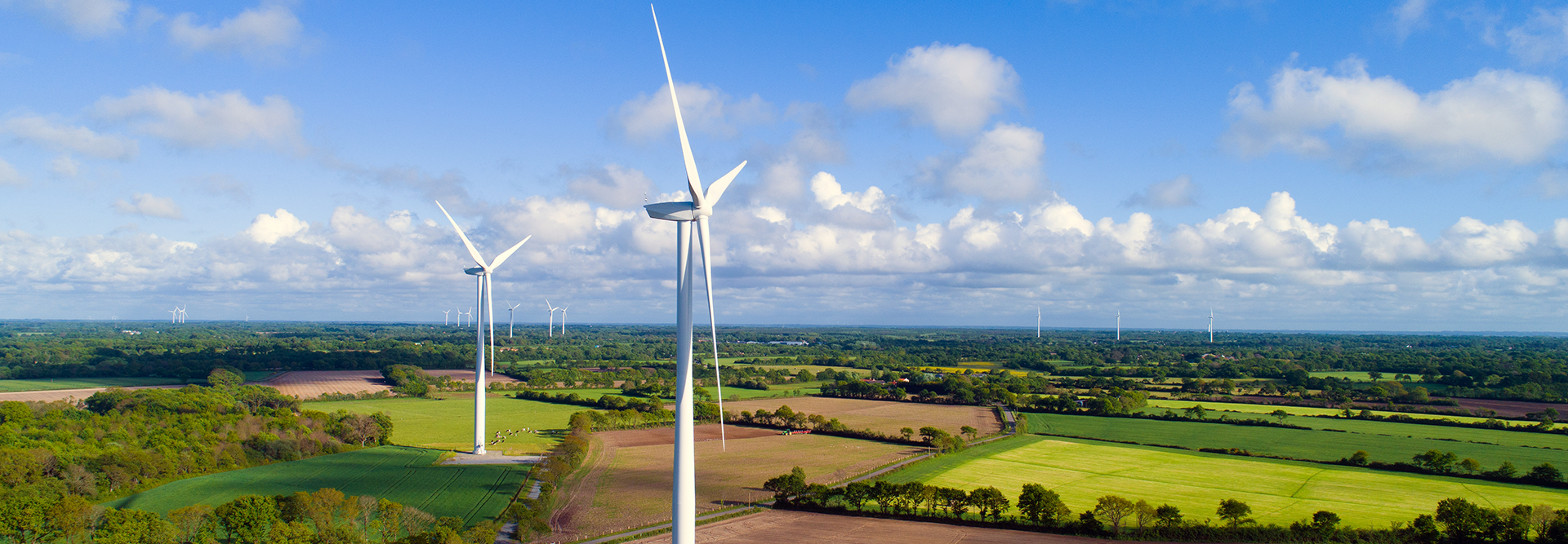 Das Bild zeigt eine Landschaft mit mehreren Windkraftanlagen auf freiem Feld. Diese Turbinen werden zur Erzeugung von erneuerbarer Energie durch Windkraft genutzt. Die Umgebung ist grün und ländlich mit einigen Ackerflächen. Es gibt einen blauen Himmel mit ein paar Wolken.