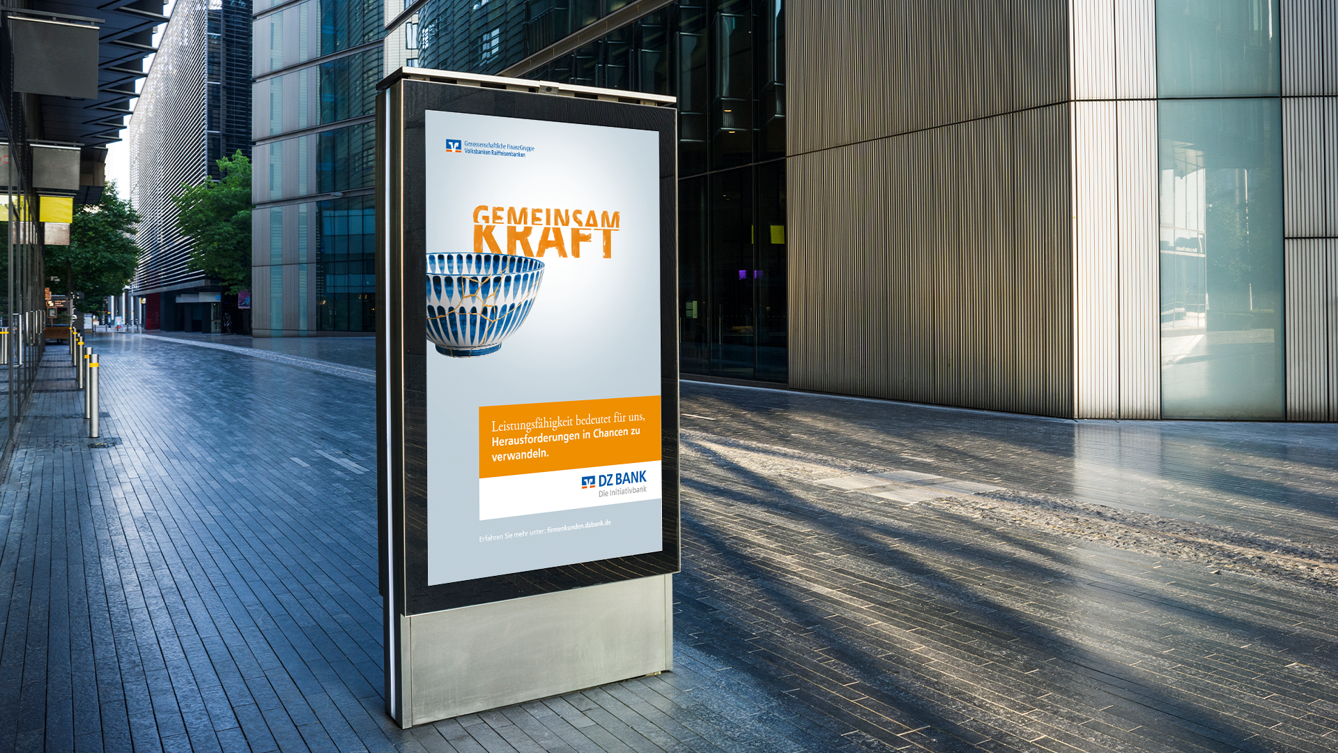 Das Bild zeigt ein Werbeposter eines Finanzinstituts, in dem das Konzept von "Gemeinsam Kraft" hervorgehoben wird. Auf dem Poster ist eine stilisierte Darstellung einer Gruppe von Menschen oder einem ähnlich geformten Objekt zu sehen, was möglicherweise den Zusammenhalt oder das Gemeinschaftsgefühl symbolisieren soll. Der Text und der Slogan, die in oranger und blauer Schrift gehalten sind, scheinen ein Gefühl der Verbundenheit und Kraft in Zahlen zu vermitteln, das Kernthema der Werbung umspannt. Das Werbeposter befindet sich in einer modernen, urbanen Umgebung, vermutlich vor einem Bürogebäude oder einer Fußgängerzone, und zielt darauf ab, Passanten oder die breite Öffentlichkeit anzusprechen.