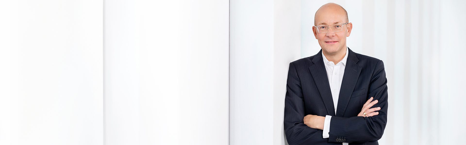 Die Aufnahme zeigt eine Person in einem dunklen Anzug mit gekreuzten Armen vor einem weißen Hintergrund.